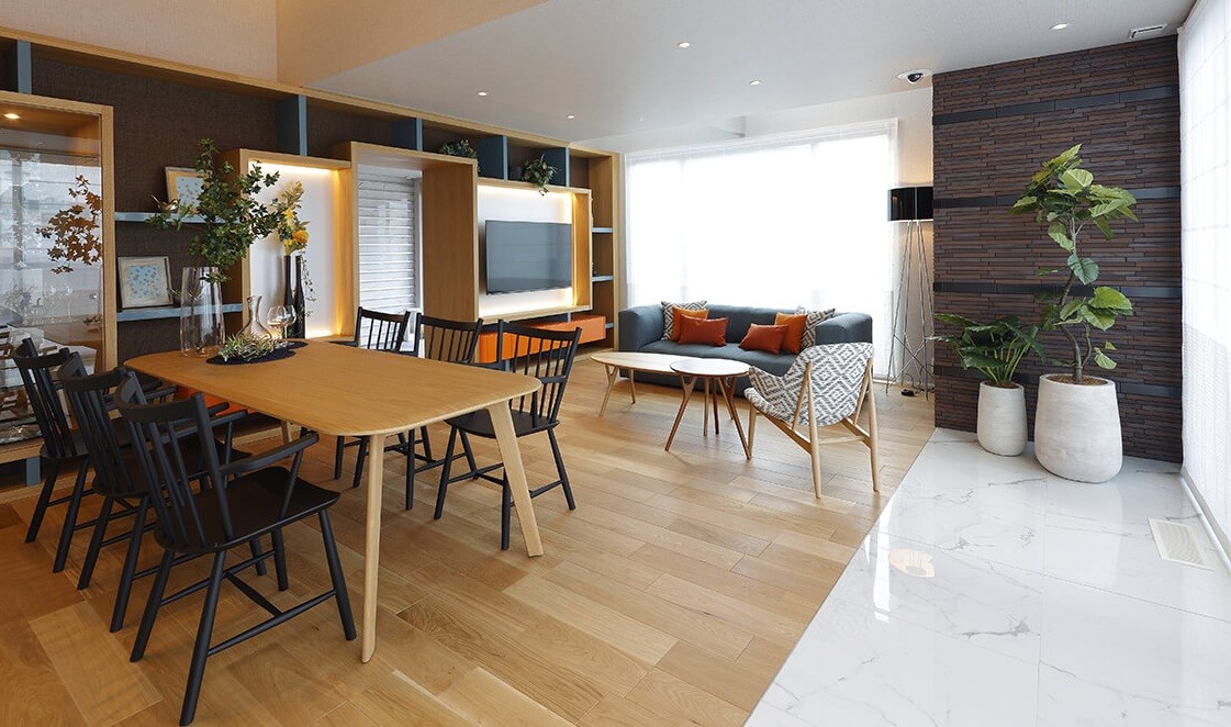 Sala de estar e jantar com mesa de madeira clara e cadeiras pretas, sof&aacute; cinza com almofadas laranja, estante com TV e plantas, e parede de tijolos escuros com vasos brancos