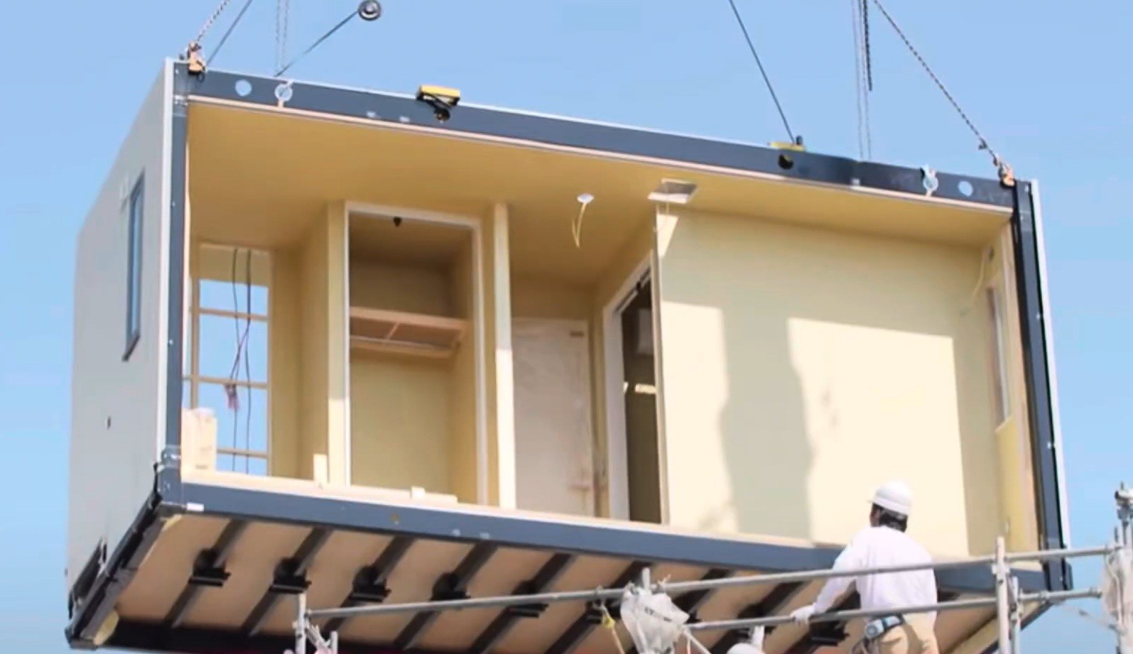Um m&oacute;dulo de casa pr&eacute;-fabricada, com paredes internas claras e janelas, sendo i&ccedil;ado por guindaste. Um trabalhador de capacete branco e camisa clara observa a opera&ccedil;&atilde;o de uma estrutura de andaimes abaixo