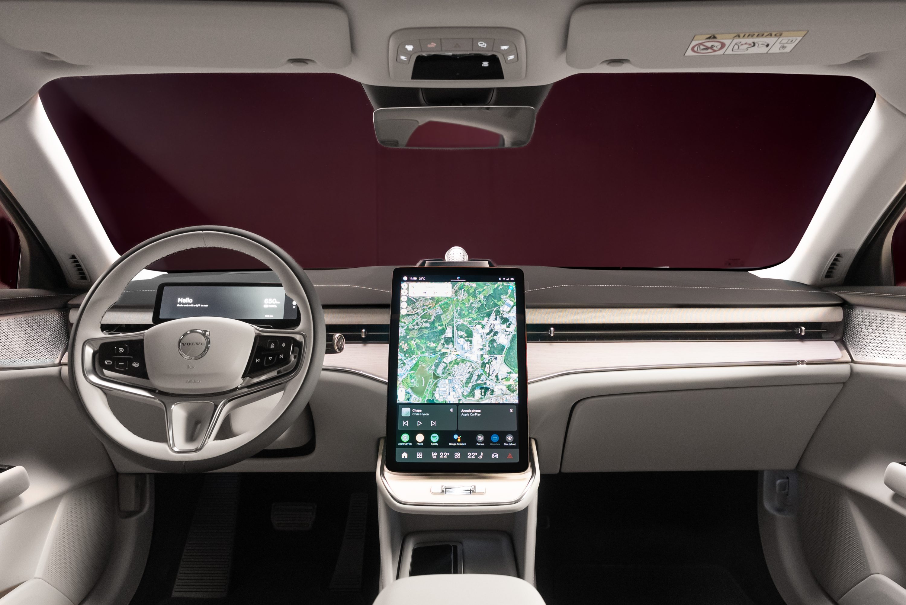 Interior de um carro moderno com volante cinza claro e tela vertical central exibindo um mapa de navega&ccedil;&atilde;o. O painel &eacute; cinza claro com detalhes em metal escovado, e o para-brisa tem um tom avermelhado