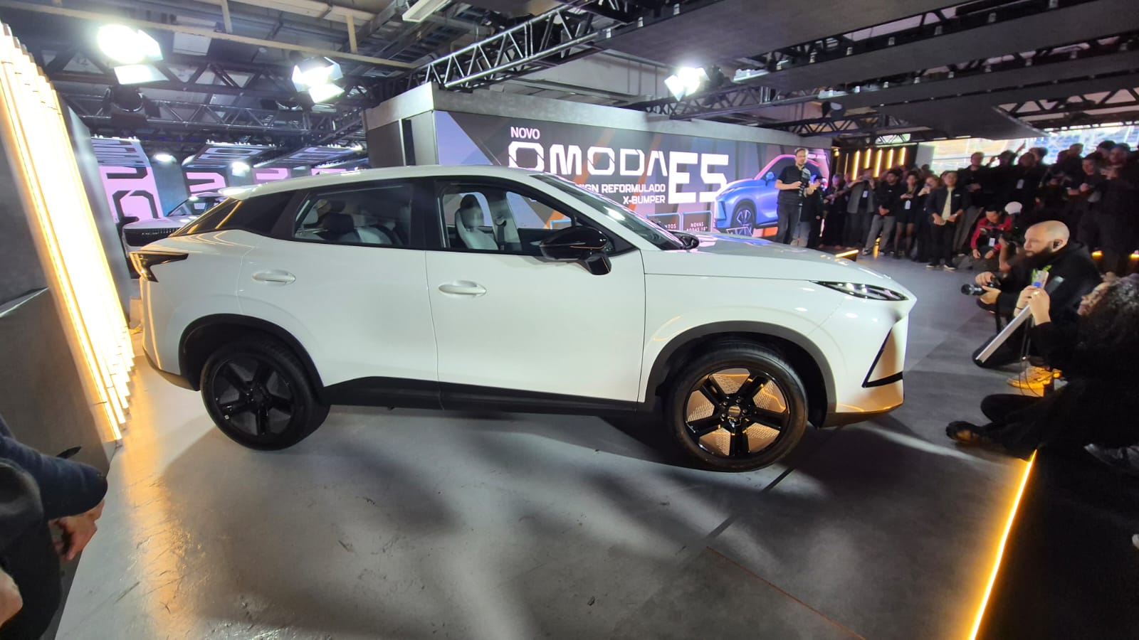SUV branco Omoda E5 em destaque num palco iluminado, com o letreiro NOVO OMODA E5 ao fundo. Pessoas e fot&oacute;grafos registram o evento.