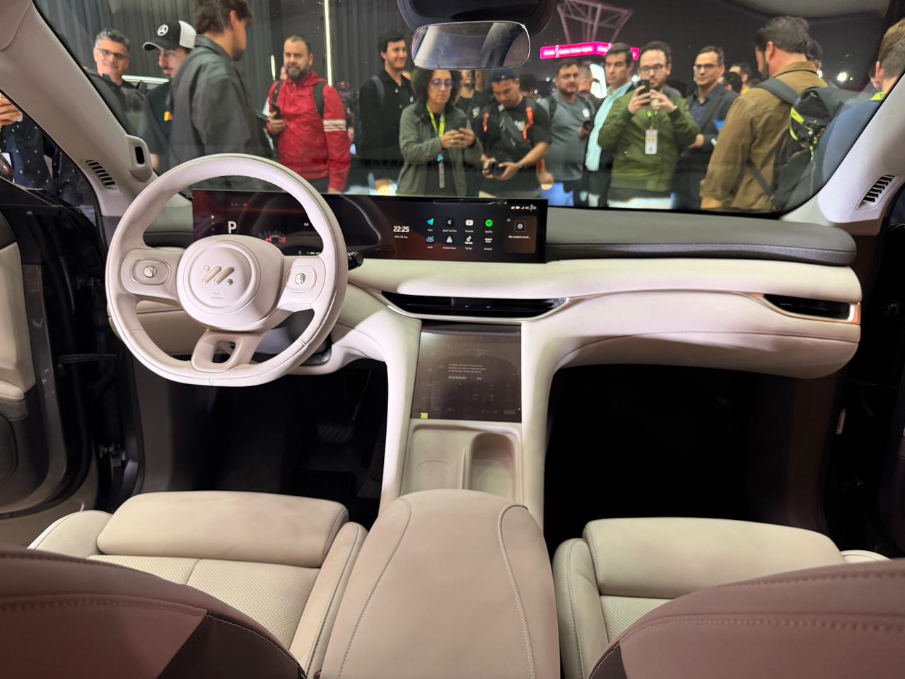 Interior de carro moderno com volante e painel brancos, bancos de couro bege. Uma tela horizontal exibe &iacute;cones de aplicativos. Ao fundo, um grupo de pessoas observa o ve&iacute;culo, algumas tirando fotos