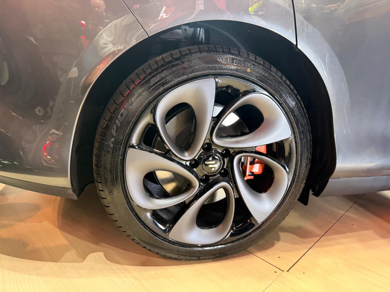 Roda de carro com design moderno, aro preto brilhante e cinco raios cinza-claro em formato de bumerangue, com pin&ccedil;a de freio laranja vis&iacute;vel ao fundo. O pneu preto tem a marca Pirelli e o carro &eacute; cinza-chumbo, refletindo luzes vermelhas. O ve&iacute;culo est&aacute; sobre um piso de madeira clara.