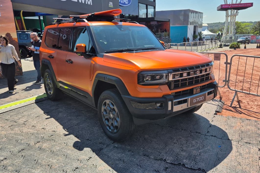 SUV laranja fosco com prancha de surf no teto, estacionado em &aacute;rea externa com piso cinza e pessoas ao fundo. O ve&iacute;culo tem grade frontal preta e cromada, far&oacute;is retangulares e para-choques robustos. &Agrave; direita, uma cerca met&aacute;lica e terreno de terra batida.