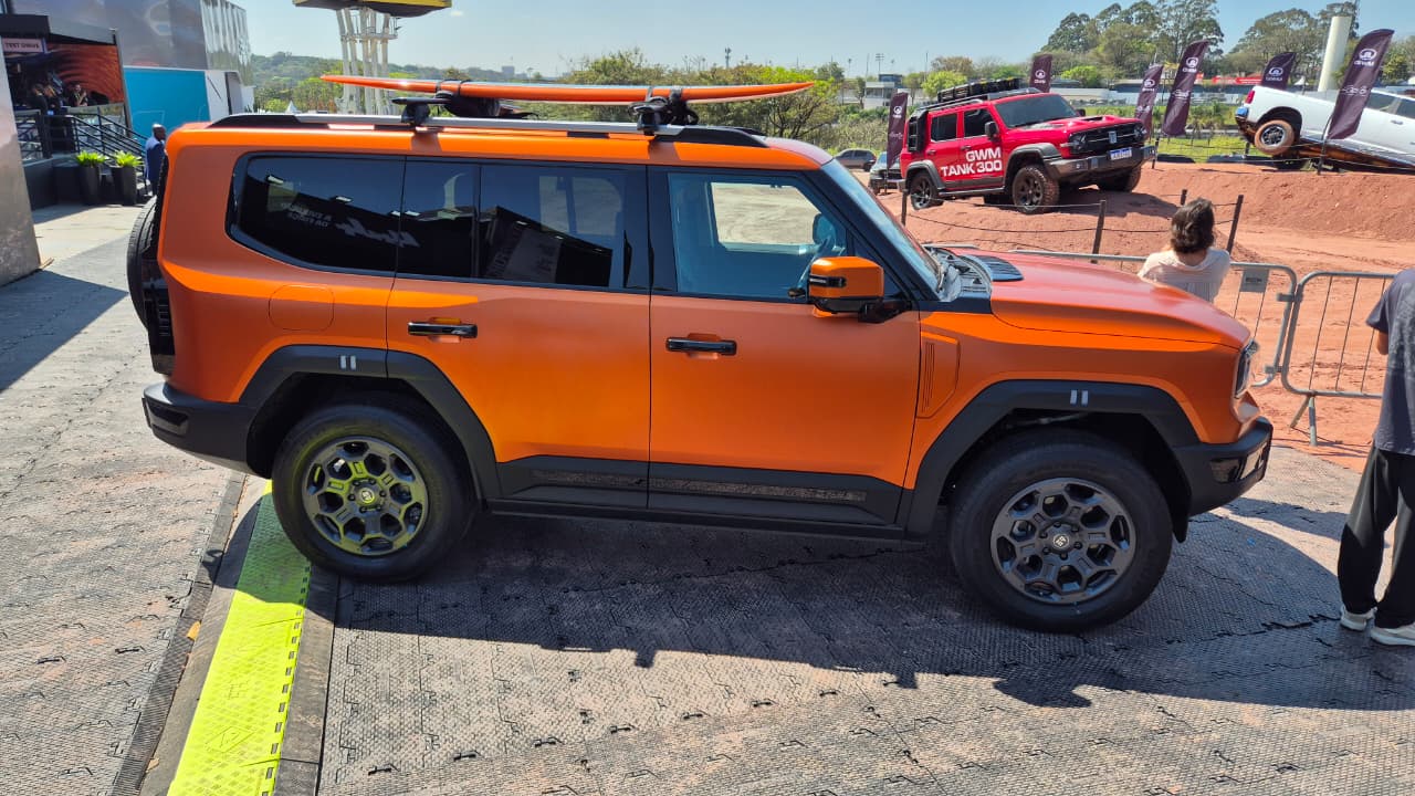 SUV laranja fosco com prancha de surf no teto, estacionado em &aacute;rea externa. Ao fundo, outros ve&iacute;culos off-road, um vermelho e um branco, em terreno acidentado, e pessoas observando. O dia est&aacute; ensolarado