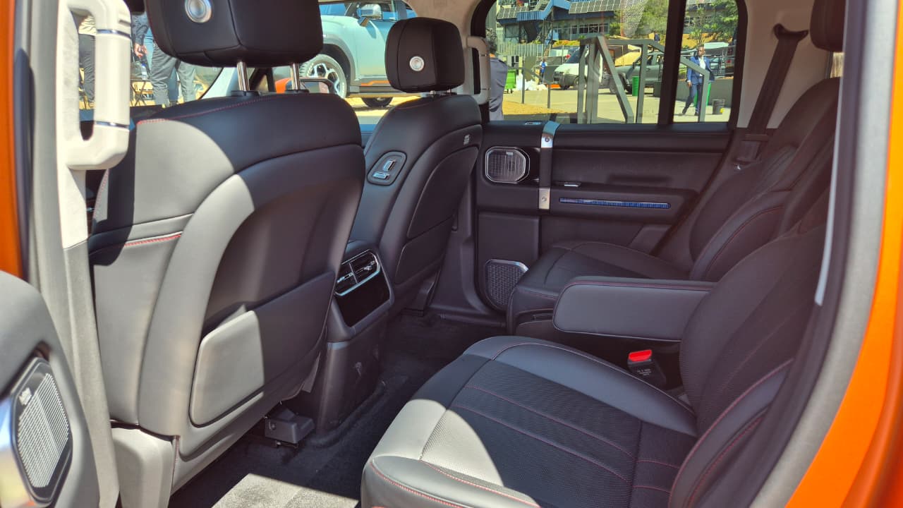 Interior de um carro laranja, mostrando os bancos traseiros pretos com detalhes em cinza e costura vermelha. H&aacute; um apoio de bra&ccedil;o central rebatido, sa&iacute;das de ar e controles no console traseiro. A janela exibe um cen&aacute;rio externo com pessoas e outros ve&iacute;culos