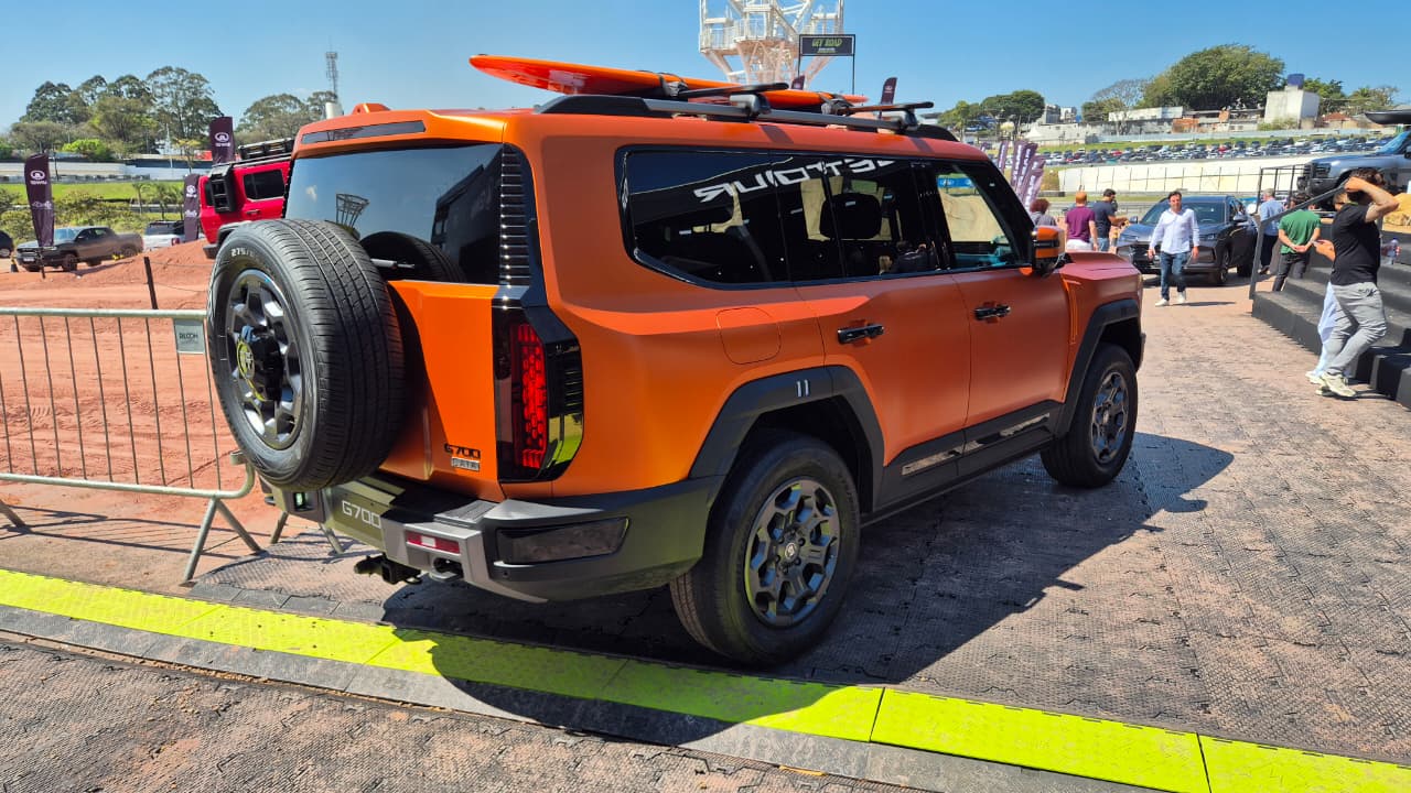 SUV laranja fosco com estepe na traseira e prancha de surfe no teto, estacionado em &aacute;rea externa com pessoas e outros ve&iacute;culos ao fundo, sob c&eacute;u azul