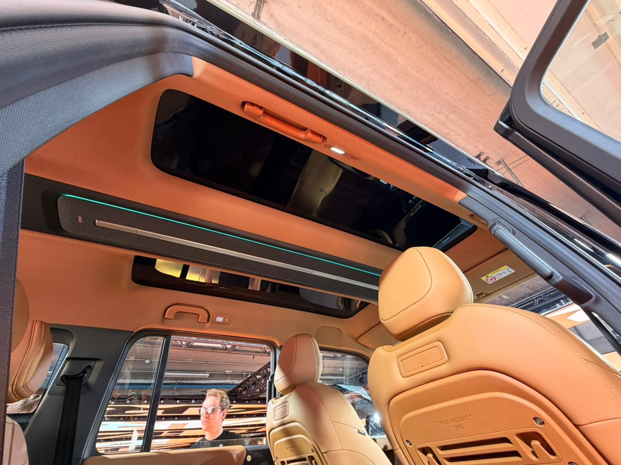 Interior de um carro com teto solar panor&acirc;mico e estofamento bege. Um homem de &oacute;culos e camiseta preta &eacute; vis&iacute;vel atrav&eacute;s da janela traseira, refletindo o ambiente externo. Detalhes de ilumina&ccedil;&atilde;o ambiente em azul e laranja no teto e al&ccedil;as de seguran&ccedil;a s&atilde;o vis&iacute;veis