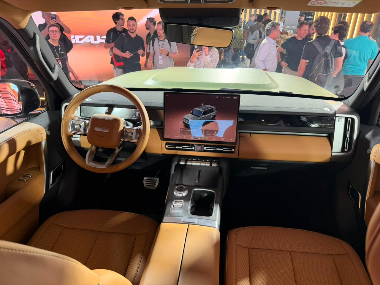 Interior de um carro moderno com volante marrom e painel em tons de bege e preto. Uma tela central exibe um SUV cinza. Pessoas em segundo plano observam o ve&iacute;culo.