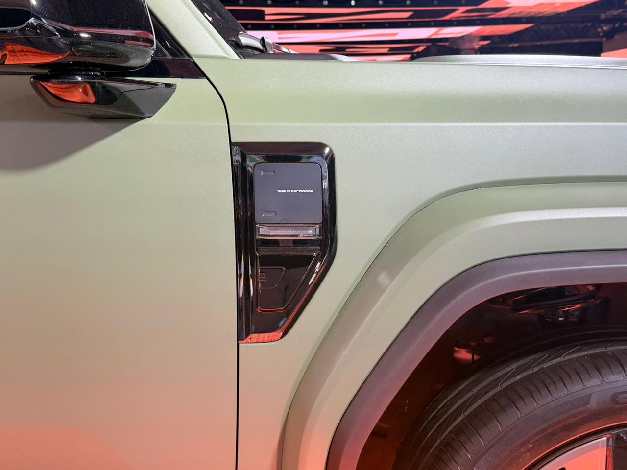 Lateral de um carro verde fosco, com retrovisor preto brilhante e um detalhe retangular escuro no para-lama dianteiro, acima da roda preta. O fundo &eacute; um borr&atilde;o laranja e preto