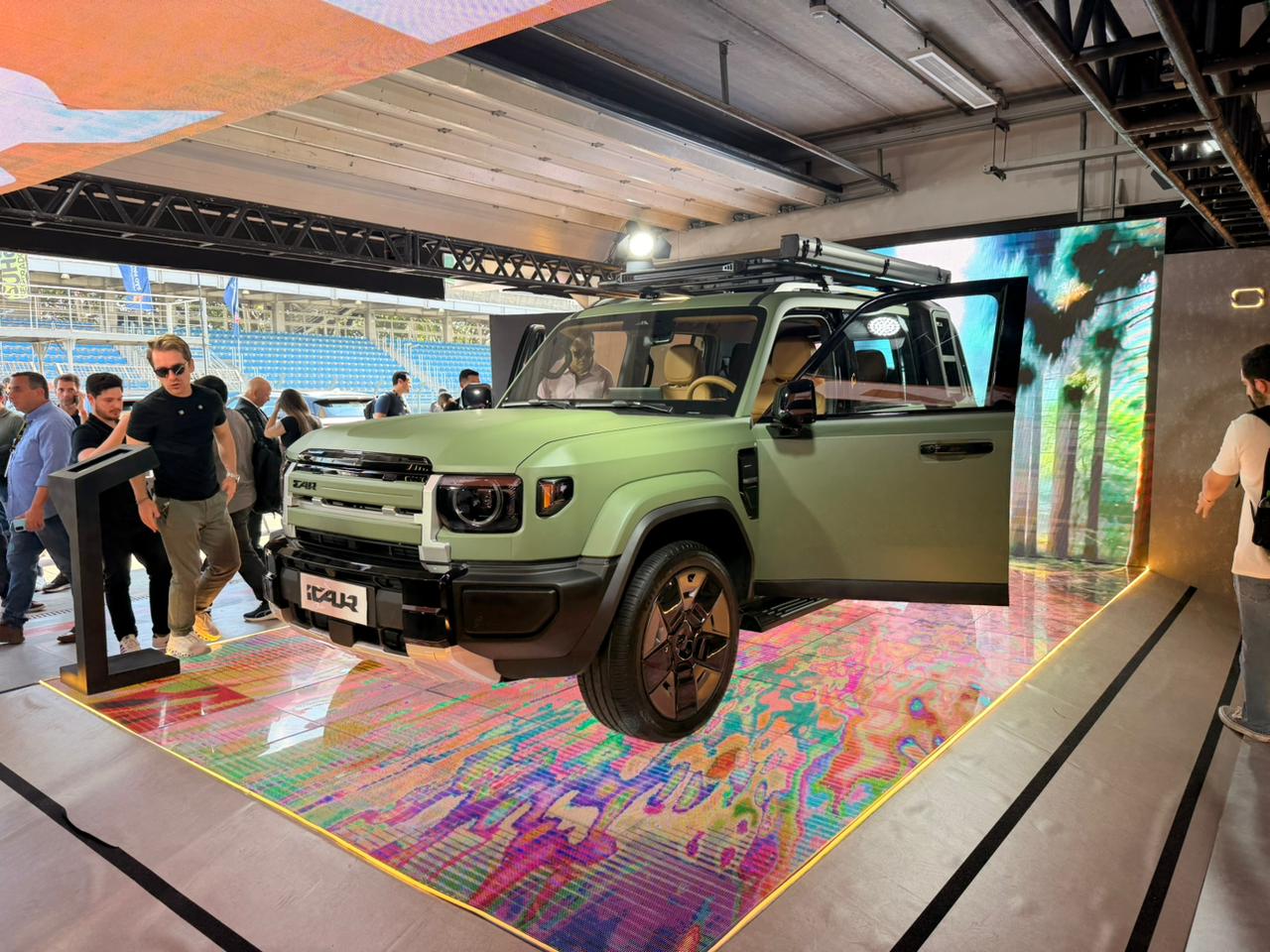 Um SUV verde-oliva com portas abertas e teto solar est&aacute; em destaque sobre um tapete colorido, com pessoas ao redor em um evento. Ao fundo, uma tela exibe palmeiras