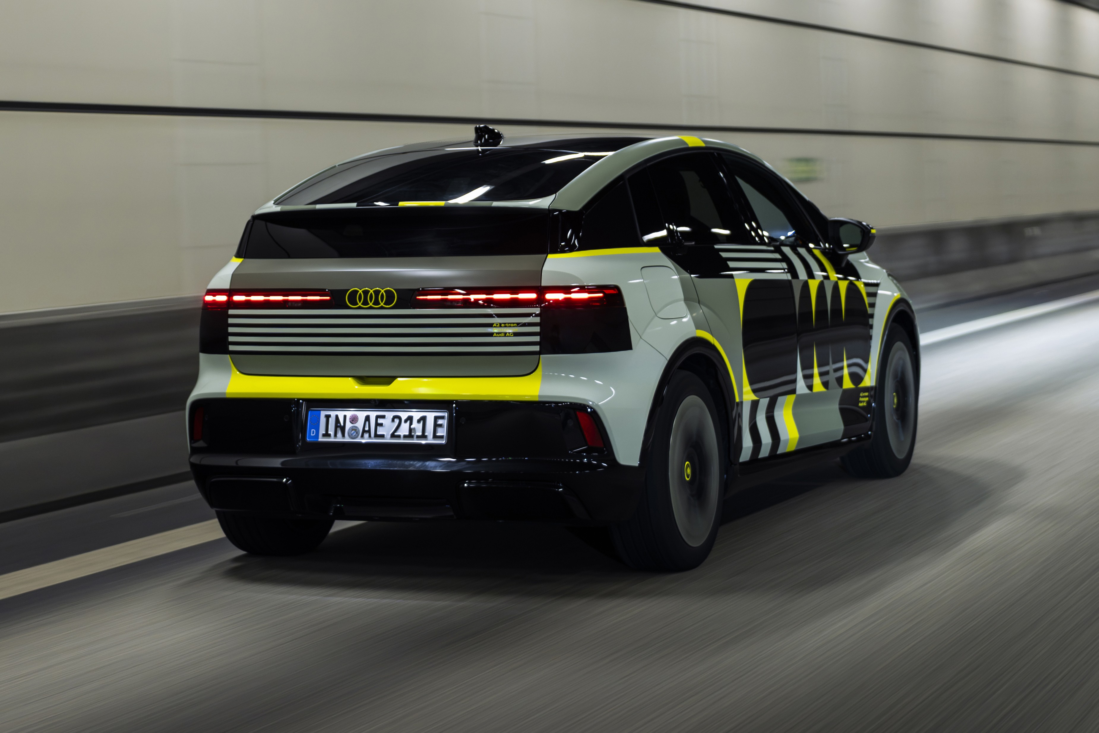 Carro Audi camuflado em cinza, preto e amarelo neon, visto por tr&aacute;s e lateralmente, em movimento r&aacute;pido dentro de um t&uacute;nel iluminado