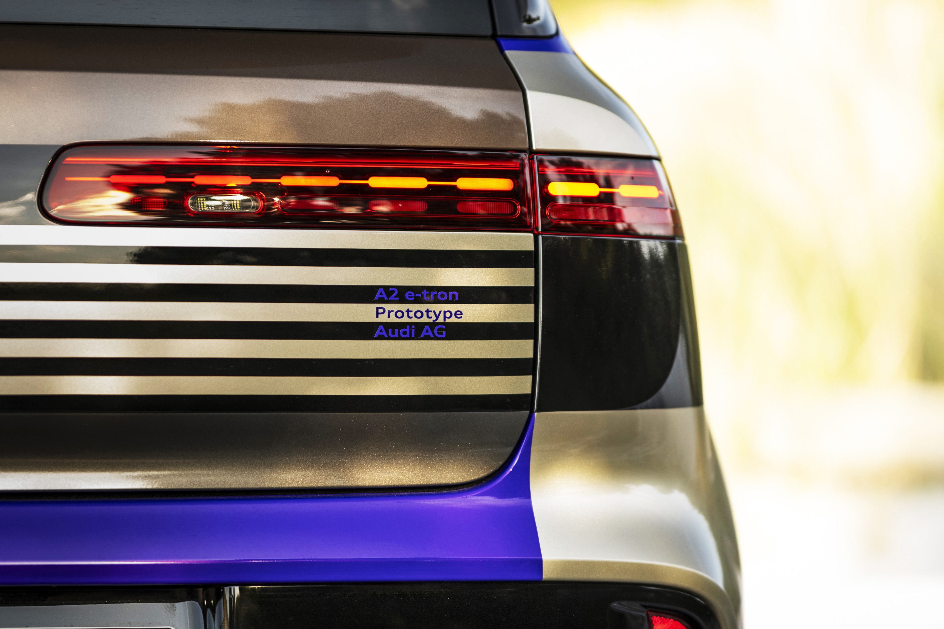 Traseira de um carro prot&oacute;tipo Audi A2 e-tron, com lanternas vermelhas e luzes de LED laranja acesas. A carroceria tem faixas horizontais pretas e douradas, com detalhes em azul vibrante na parte inferior. O fundo est&aacute; desfocado em tons claros