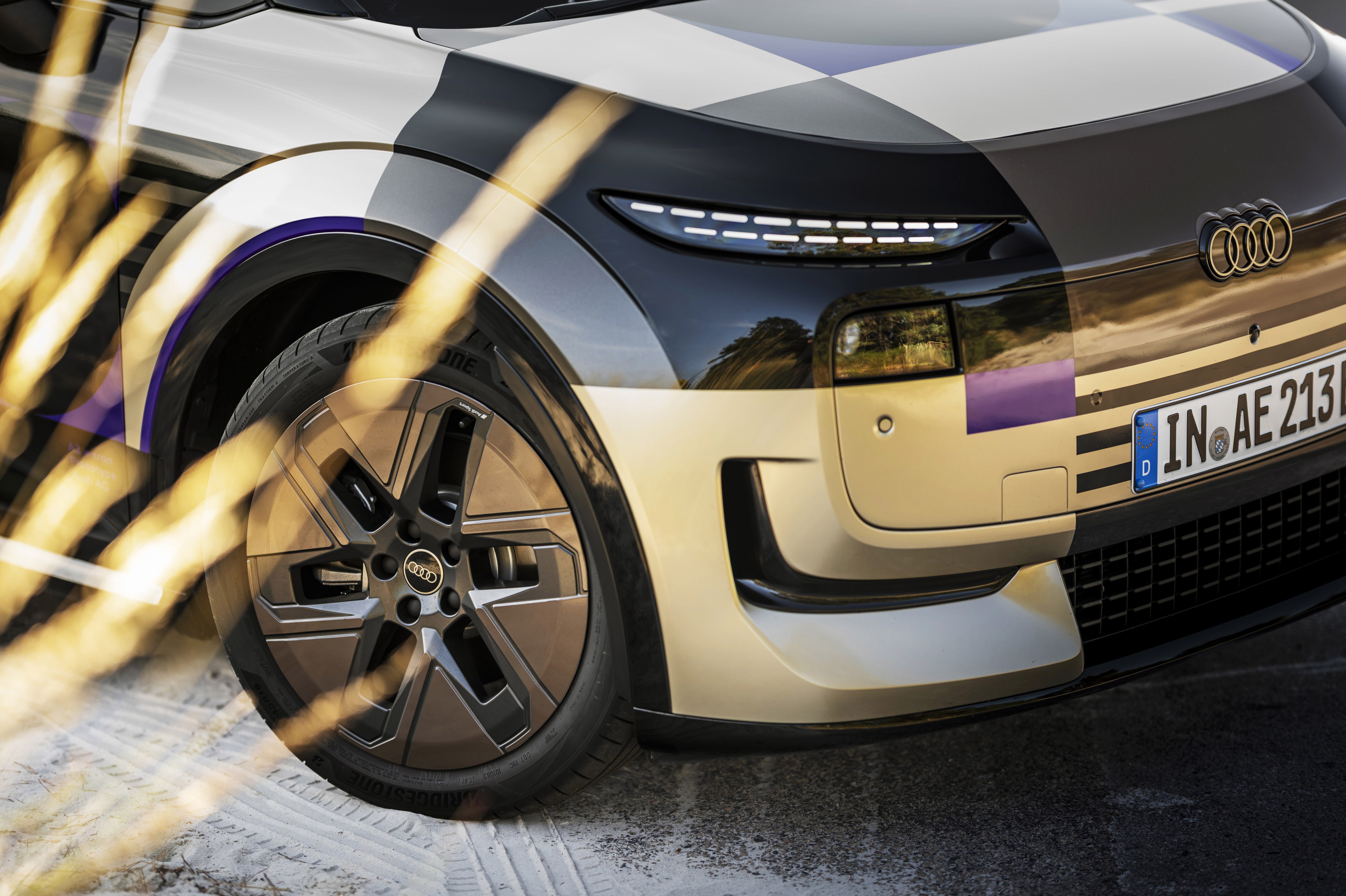 Parte frontal de um carro Audi com camuflagem quadriculada preta, branca e roxa, destacando o farol de LED, a grade e a roda com o logotipo da marca. A placa de licen&ccedil;a alem&atilde; IN AE 213 est&aacute; vis&iacute;vel. Focos de luz dourada atravessam a imagem