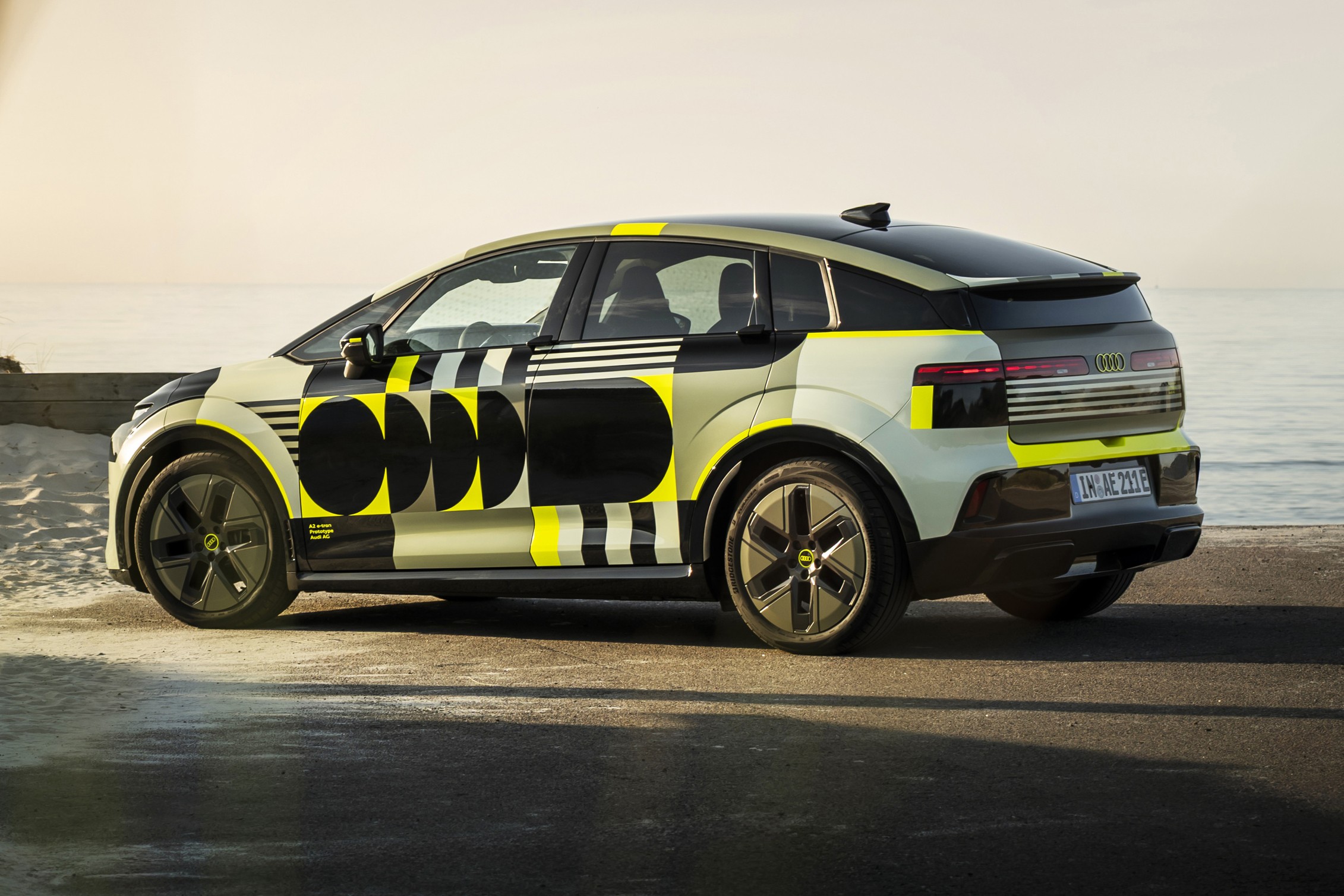 Carro el&eacute;trico camuflado com padr&otilde;es geom&eacute;tricos em preto, amarelo e cinza claro, estacionado em uma estrada de asfalto perto do mar, sob luz solar