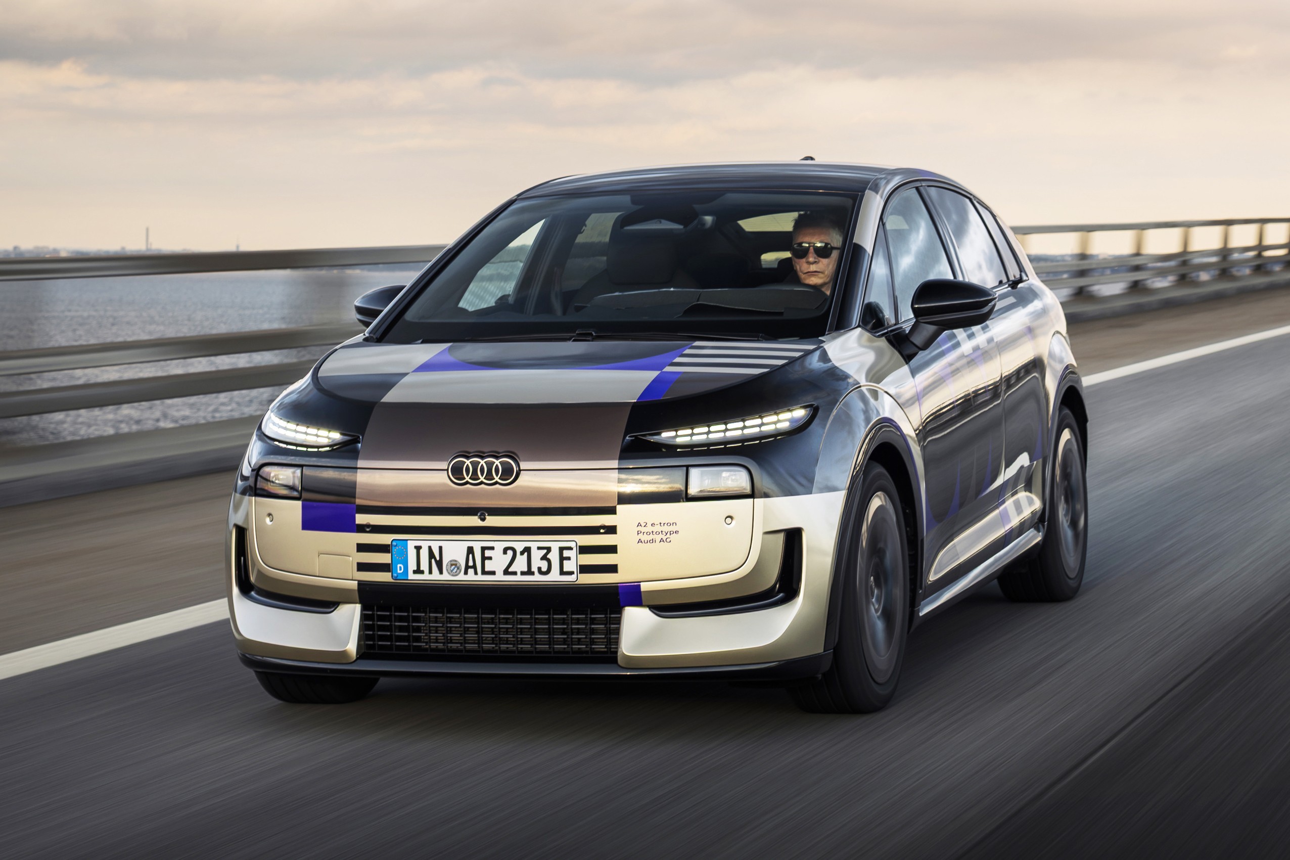 Carro Audi el&eacute;trico camuflado com padr&otilde;es geom&eacute;tricos em tons de preto, azul e bege, dirigido por uma pessoa de &oacute;culos escuros, em movimento r&aacute;pido sobre uma ponte com &aacute;gua ao fundo