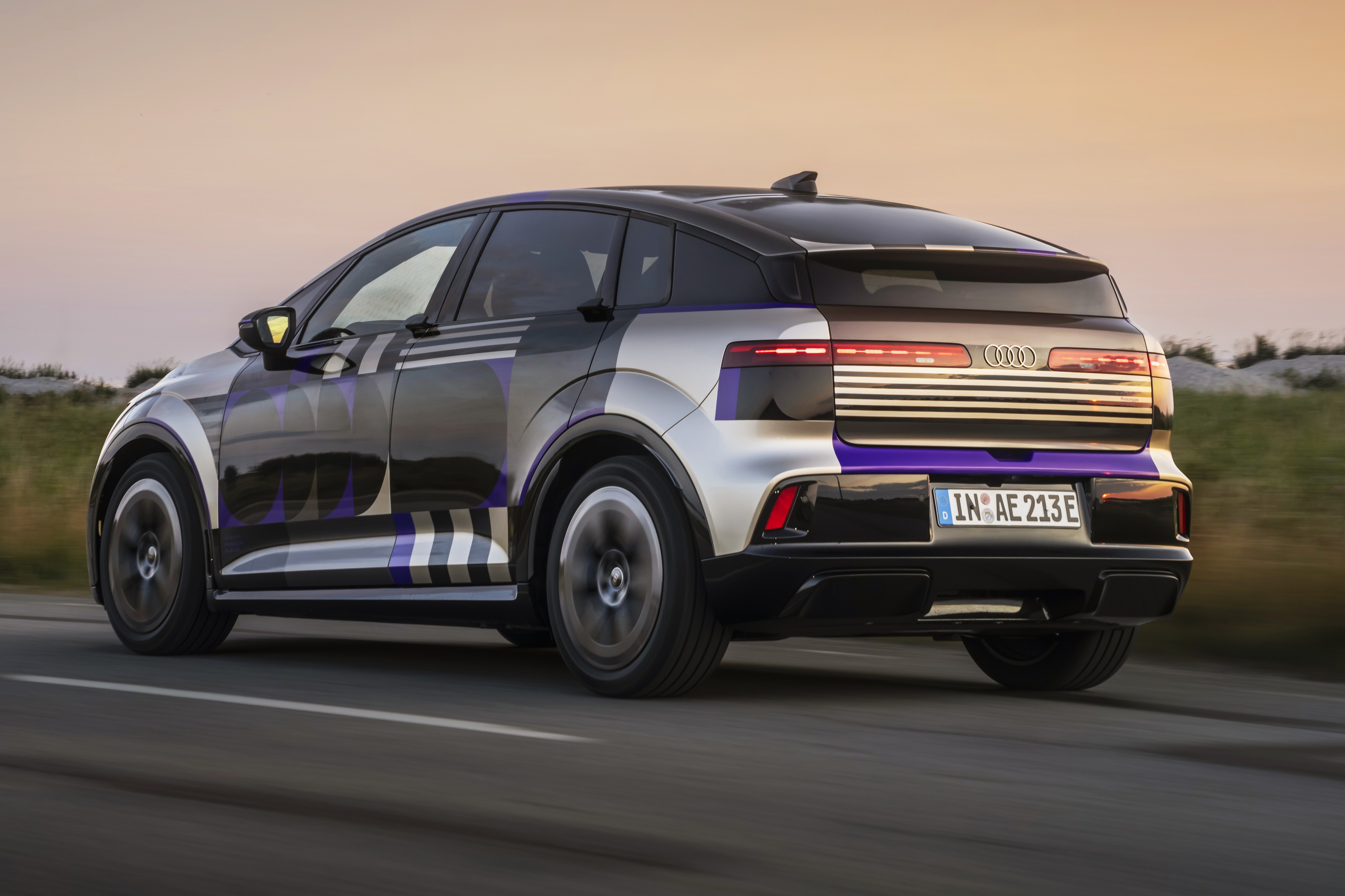 Carro Audi el&eacute;trico camuflado com padr&otilde;es geom&eacute;tricos em preto, prata e roxo, visto de traseira lateral, em movimento numa estrada, com c&eacute;u claro ao fundo