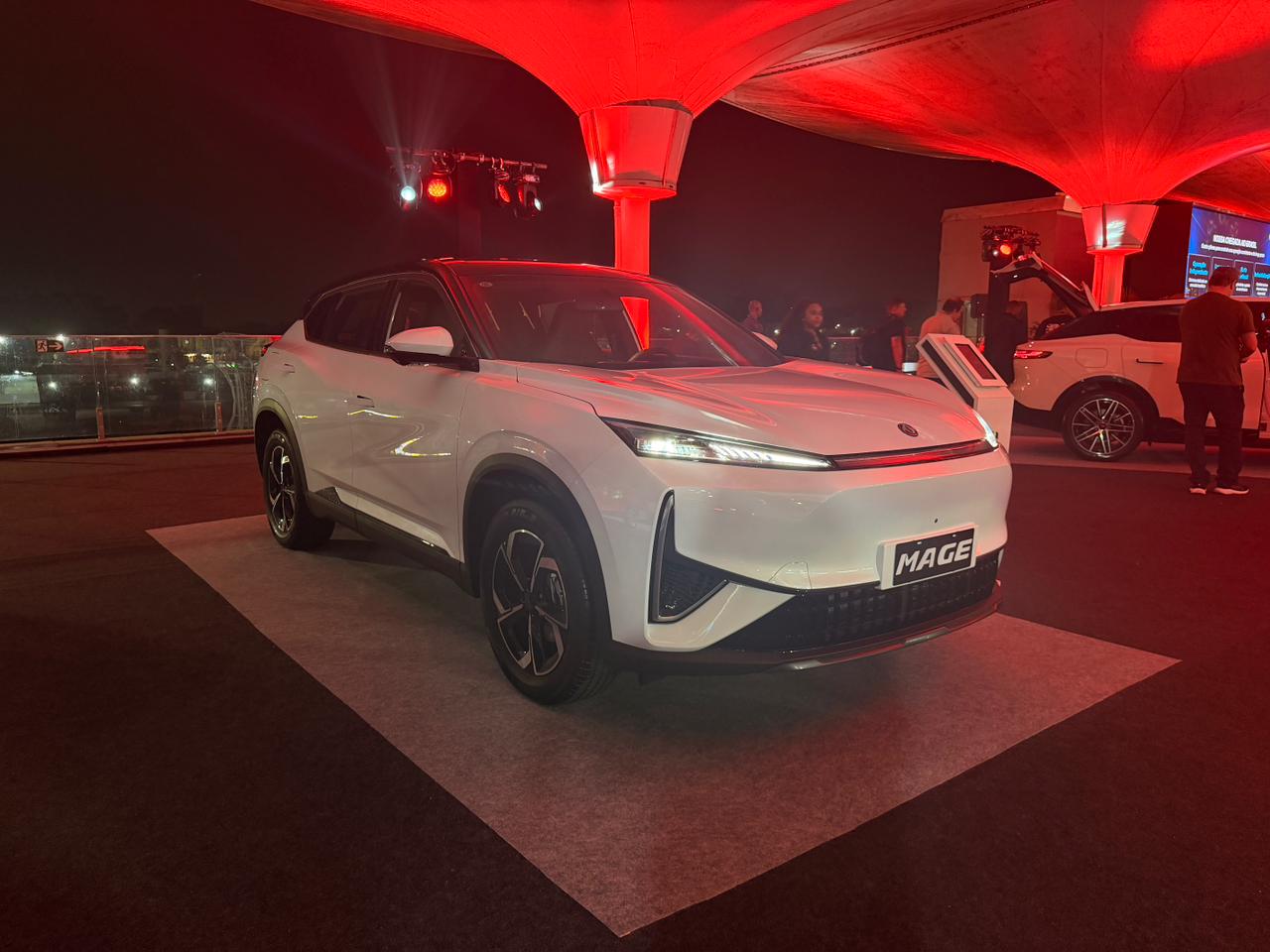 Carro SUV branco moderno, modelo Mage, em destaque sob ilumina&ccedil;&atilde;o vermelha em evento noturno. Outro carro branco e pessoas ao fundo.