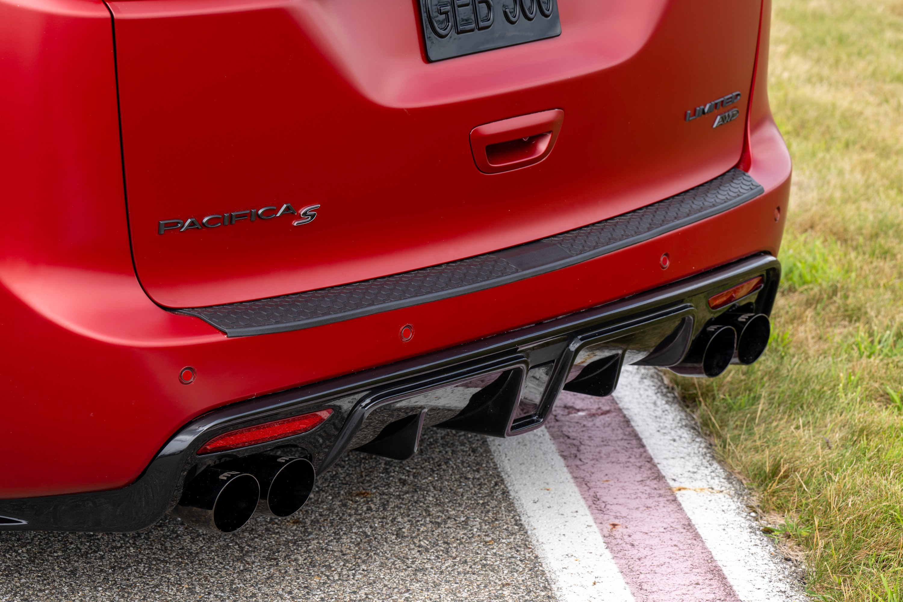 Traseira de um carro vermelho com o nome PACIFICA S em relevo. Possui um para-choque preto texturizado, difusor traseiro preto com quatro sa&iacute;das de escapamento e detalhes vermelhos nas luzes. O ve&iacute;culo est&aacute; em uma pista de asfalto com faixas brancas e vermelhas, e grama verde ao lado