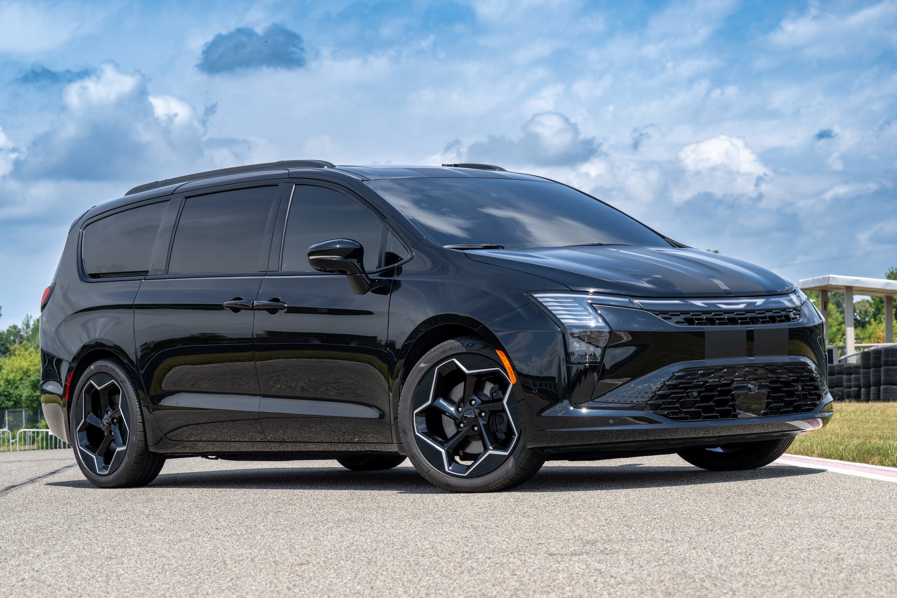 Minivan Chrysler Pacifica preta com vidros escuros e rodas esportivas pretas e brancas, estacionada em uma pista de asfalto sob c&eacute;u azul com nuvens esparsas