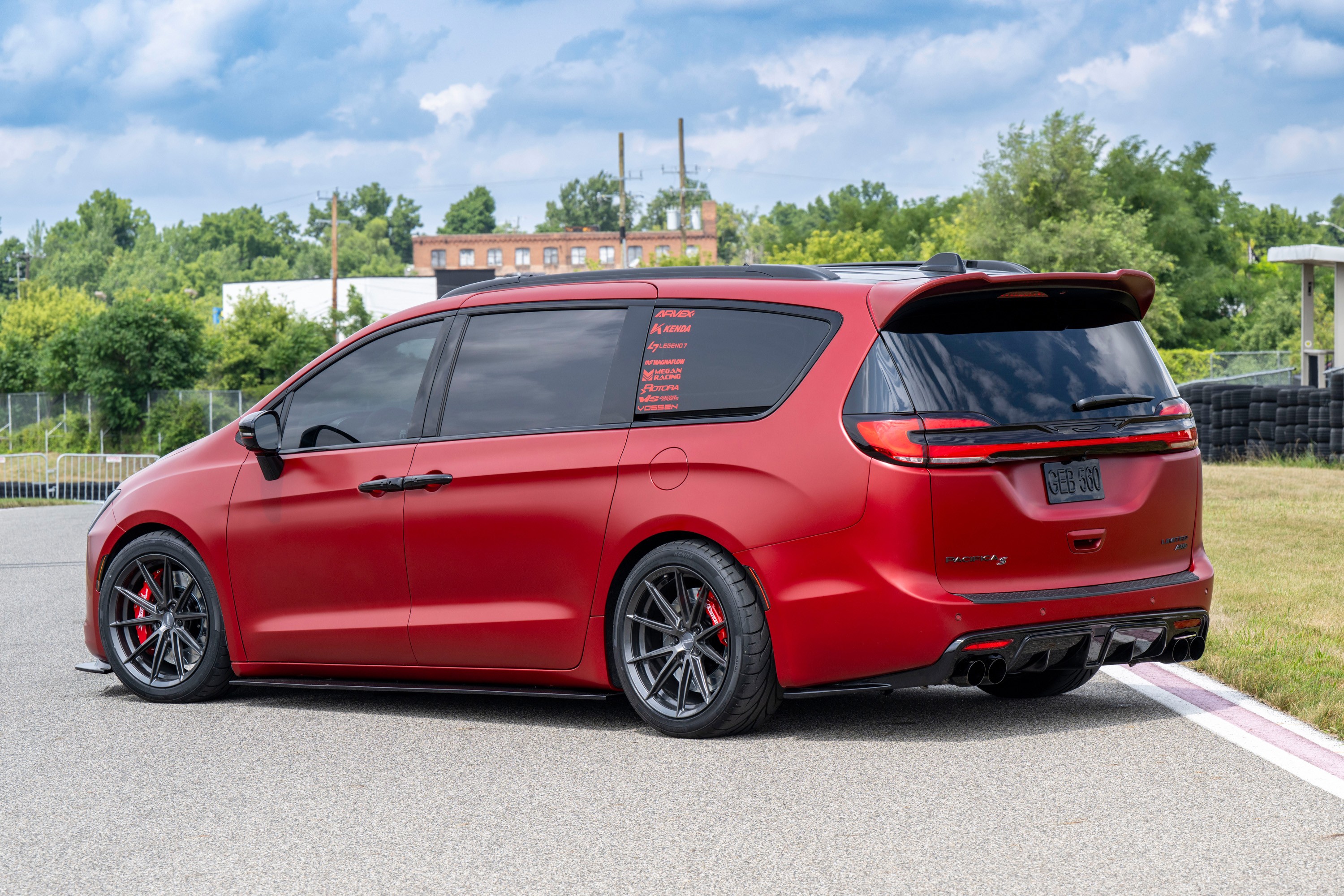 Minivan vermelha fosca, com rodas esportivas pretas e pin&ccedil;as de freio vermelhas, estacionada em uma pista de corrida. O ve&iacute;culo tem vidros escuros e adesivos na janela traseira. Ao fundo, &aacute;rvores, um pr&eacute;dio de tijolos e um c&eacute;u azul com nuvens brancas