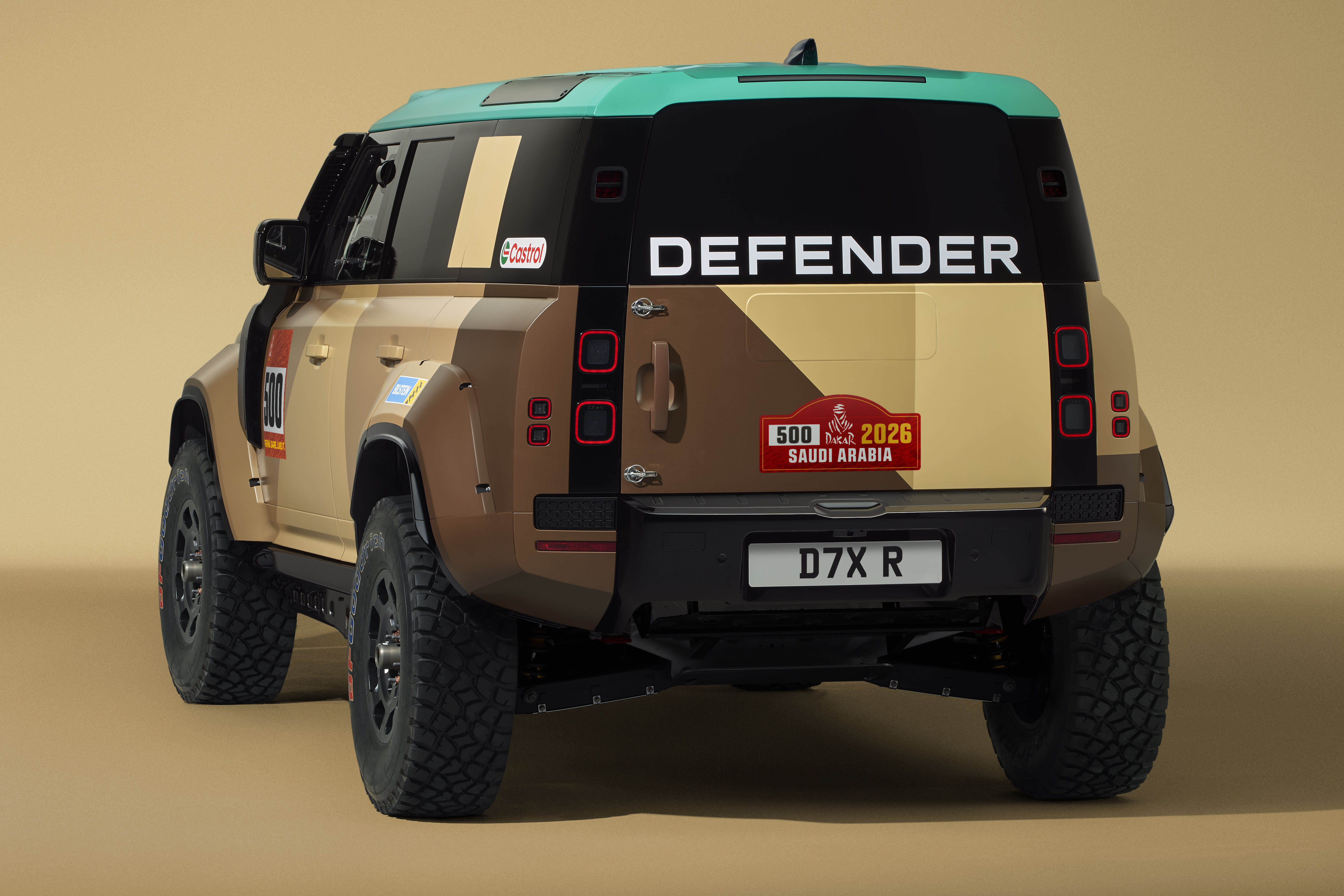 Traseira de um Land Rover Defender com pintura camuflada em tons de bege e marrom, teto verde-&aacute;gua. O nome DEFENDER est&aacute; em branco na parte superior da porta traseira. H&aacute; um adesivo vermelho com 500 2026 SAUDI ARABIA e a placa D7X R na parte inferior. O ve&iacute;culo possui pneus robustos para off-road