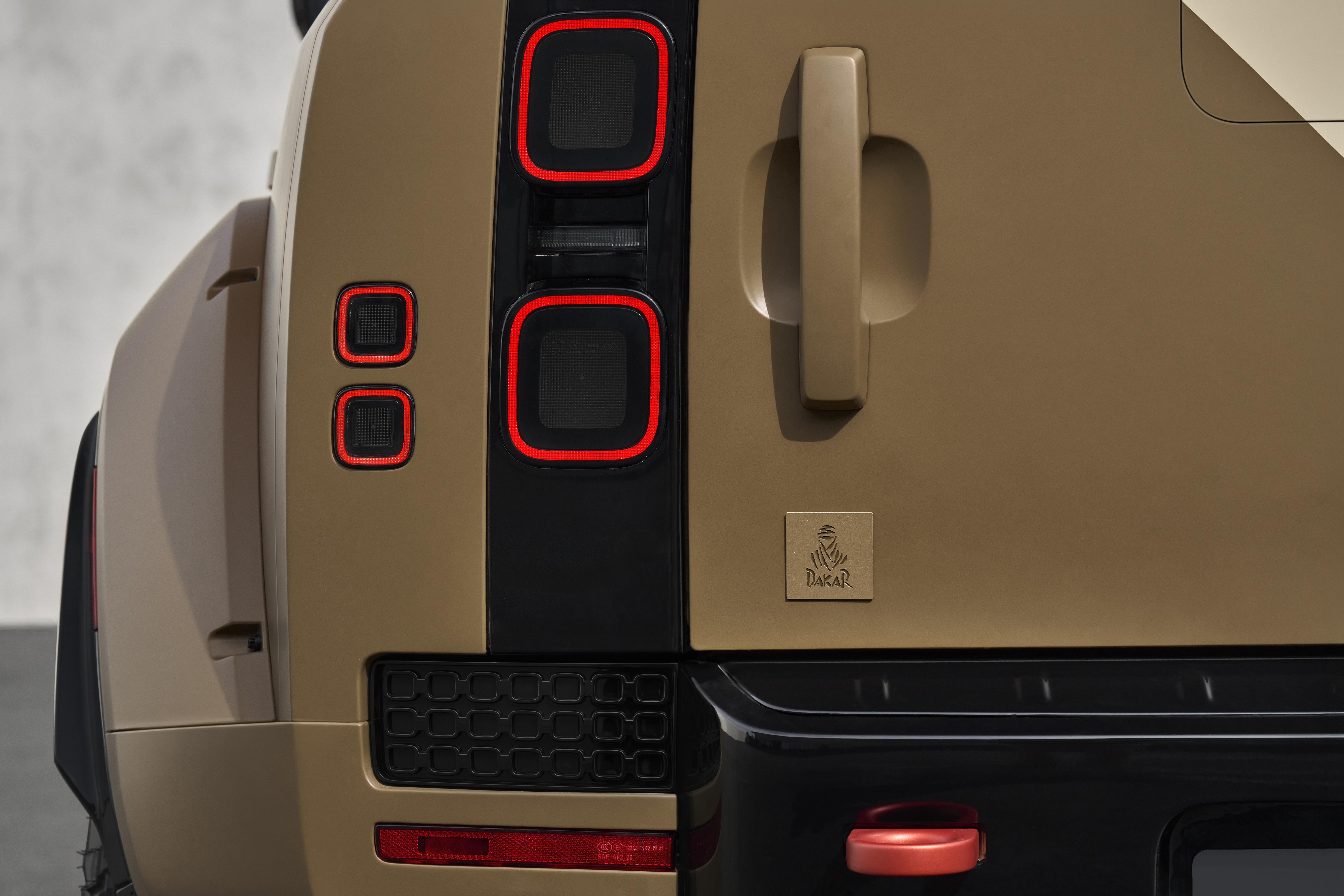 Traseira de um ve&iacute;culo utilit&aacute;rio esportivo cor areia, com lanternas quadradas de contorno vermelho, ma&ccedil;aneta da porta traseira e emblema Dakar dourados, e para-choque preto