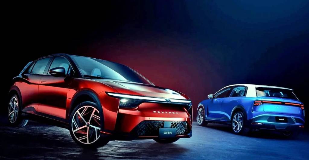 Dois carros el&eacute;tricos modernos em um est&uacute;dio escuro: um SUV vermelho-escuro em primeiro plano, com far&oacute;is acesos e a marca FOXTRO na grade, e um SUV azul com teto branco ao fundo, visto de traseira