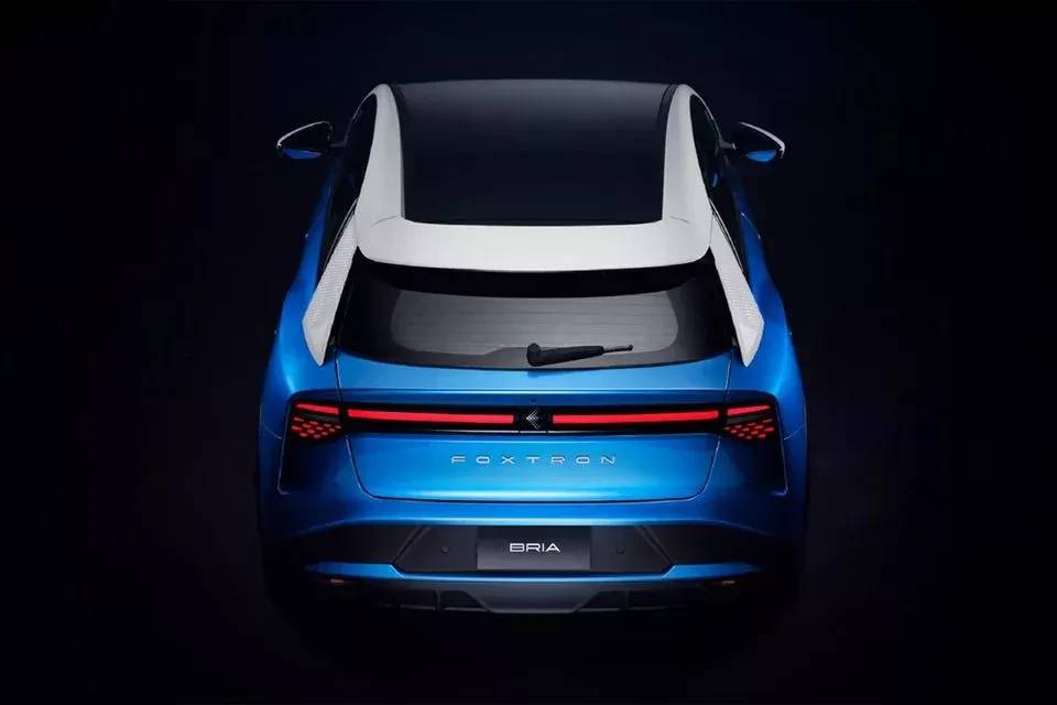 Traseira de um carro el&eacute;trico azul-met&aacute;lico, modelo Foxtron Bria, com teto preto e detalhes brancos nas laterais. As lanternas traseiras s&atilde;o uma faixa horizontal vermelha que se estende por toda a largura do ve&iacute;culo, com padr&otilde;es de pontos luminosos nas extremidades. O limpador de para-brisa est&aacute; na parte superior do vidro traseiro. O carro est&aacute; em um fundo escuro, com ilumina&ccedil;&atilde;o focada na parte traseira