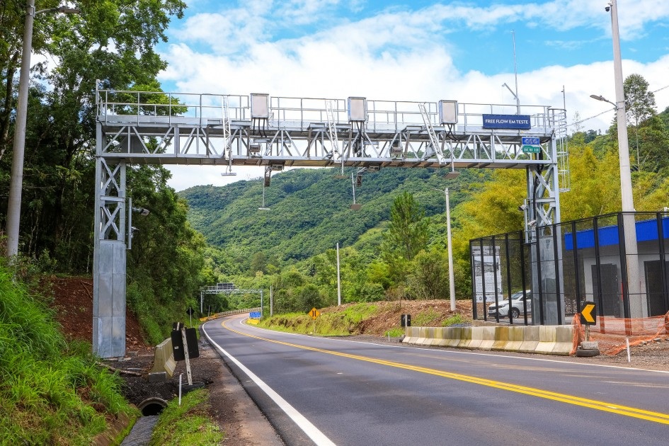 P&oacute;rtico de ped&aacute;gio free flow sobre uma rodovia sinuosa, com montanhas verdes ao fundo e c&eacute;u azul com nuvens. &Agrave; direita, uma estrutura azul e preta cercada por grades.
