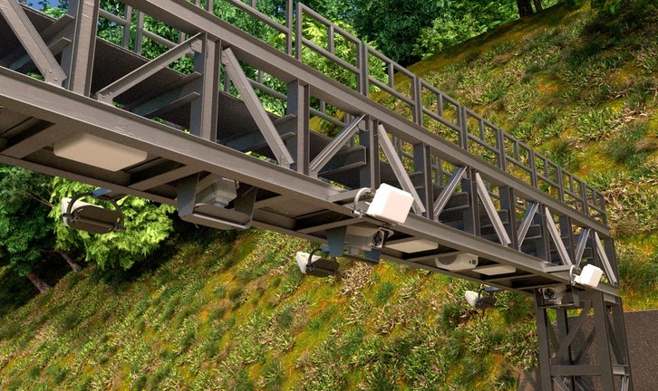 Estrutura met&aacute;lica de uma passarela ou p&oacute;rtico de ped&aacute;gio eletr&ocirc;nico, com c&acirc;meras e sensores instalados na parte inferior, sobre uma estrada escura. Ao fundo, um morro coberto por vegeta&ccedil;&atilde;o verde e &aacute;rvores