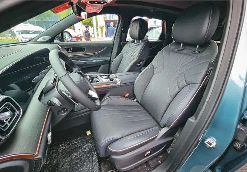 Interior de um carro moderno com bancos de couro preto e costura em losango, volante multifuncional e painel escuro. O banco do motorista est&aacute; em primeiro plano, com detalhes em costura clara e ajuste el&eacute;trico. Ao fundo, pessoas e um barco branco s&atilde;o vis&iacute;veis atrav&eacute;s dos vidros