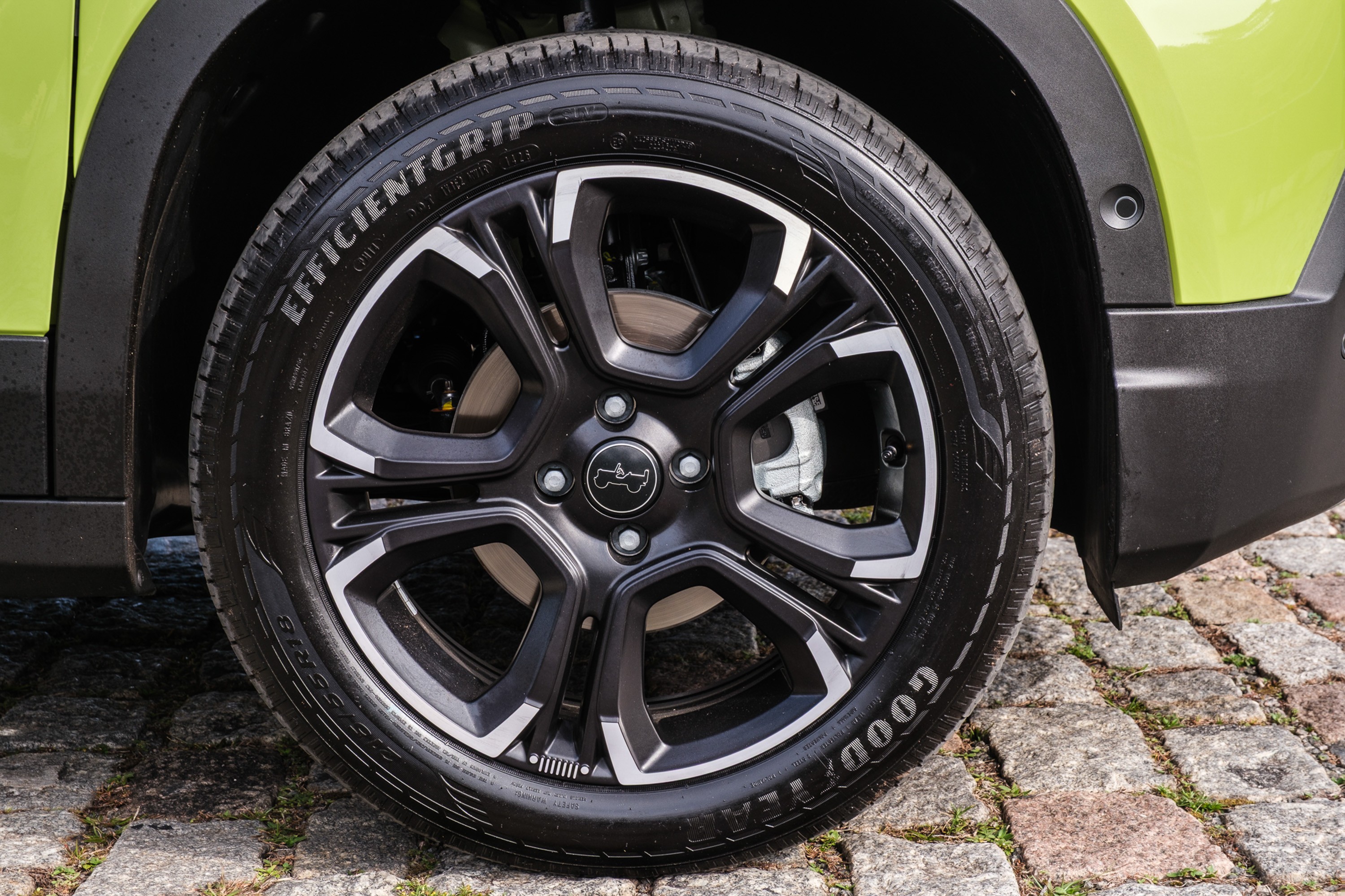 Pneu Goodyear EfficientGrip em roda preta com detalhes prateados, parte de um carro verde-lim&atilde;o, estacionado sobre paralelep&iacute;pedos