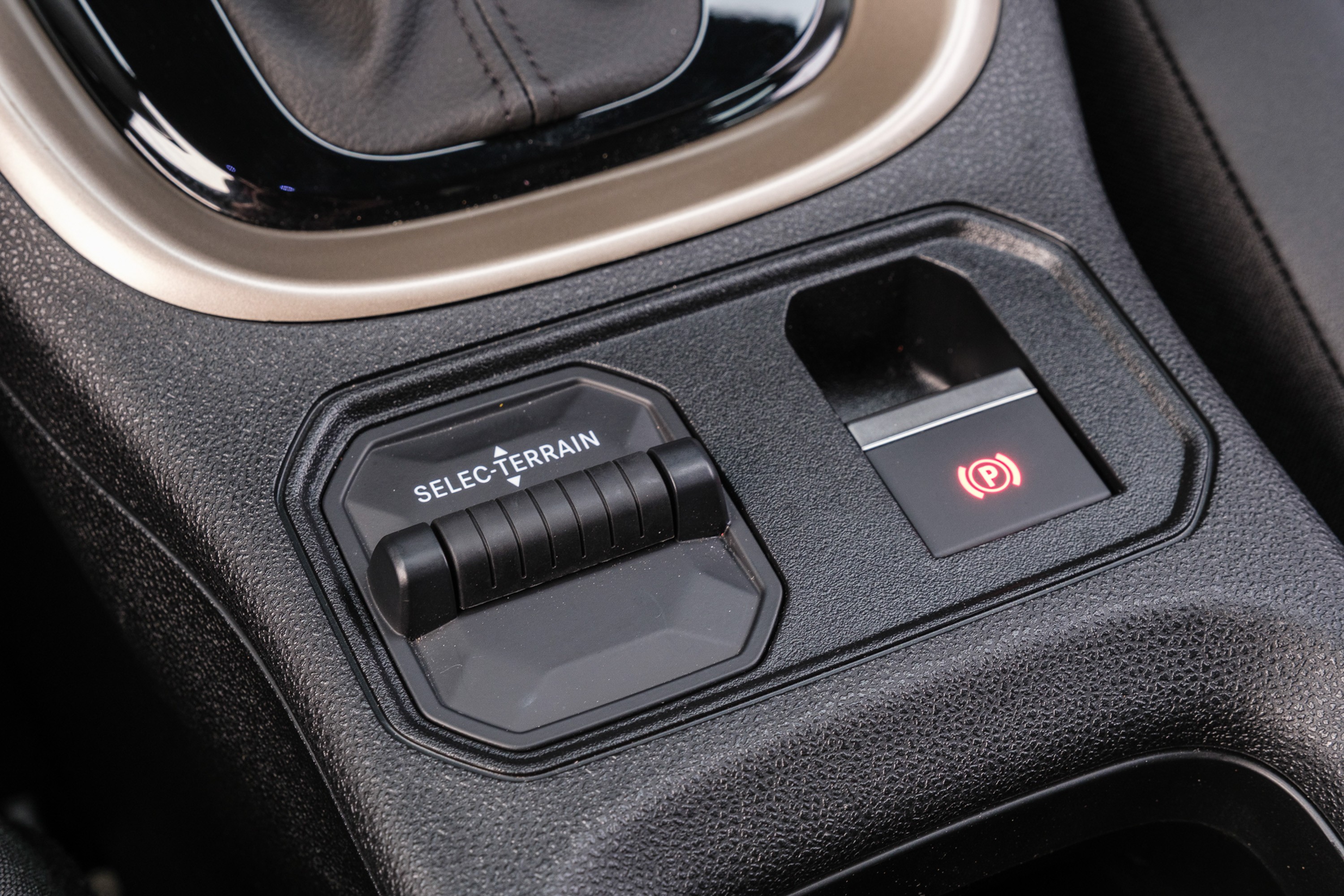 Detalhe do console central de um carro, mostrando o seletor de terreno SELEC-TERRAIN e o bot&atilde;o do freio de estacionamento eletr&ocirc;nico com a luz vermelha acesa
