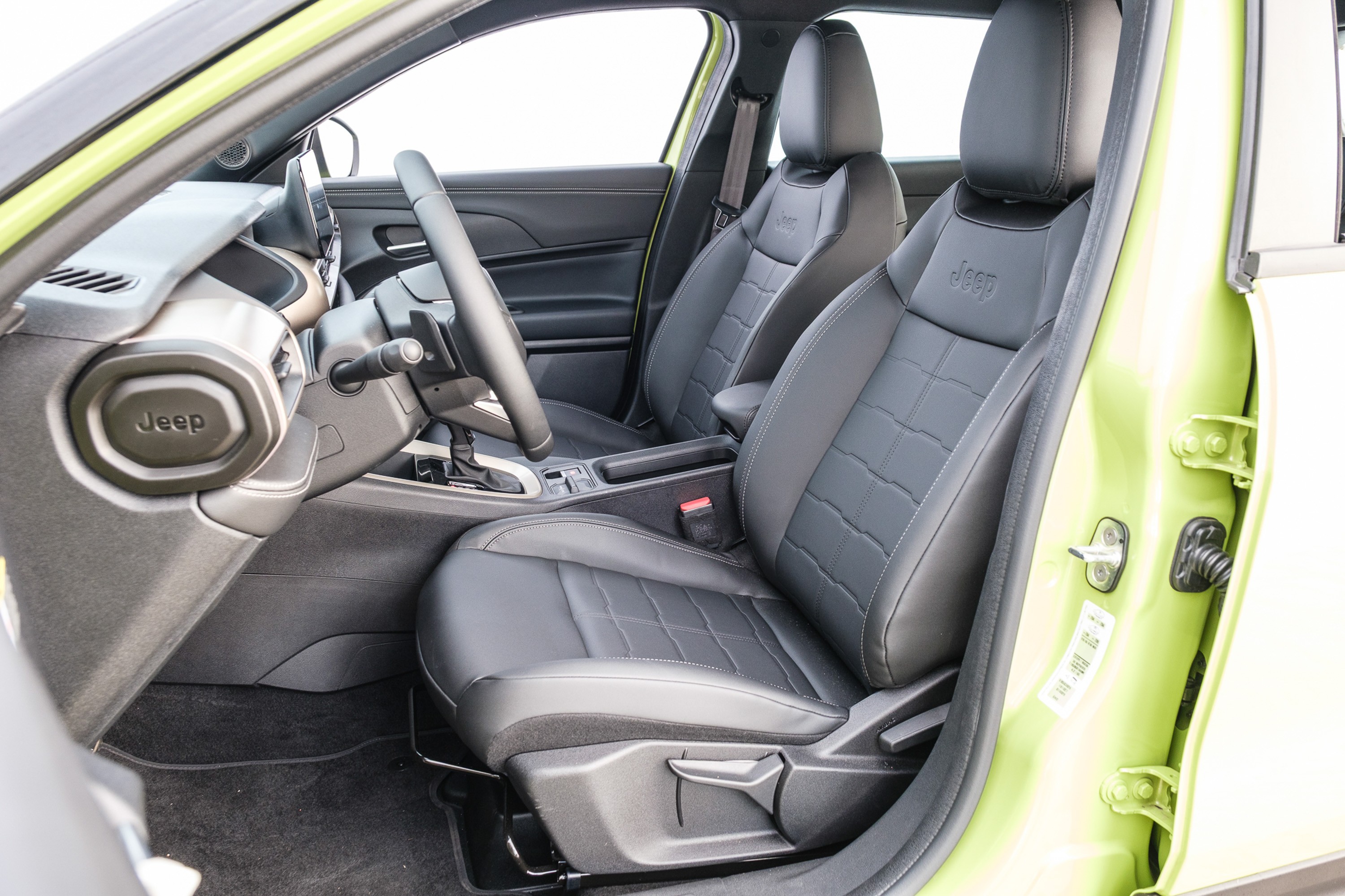 Interior de um carro Jeep verde-lim&atilde;o, com bancos de couro preto e detalhes em cinza. O volante e o painel s&atilde;o vis&iacute;veis, com o logotipo Jeep no painel. A porta do motorista est&aacute; aberta, revelando a cor vibrante do ve&iacute;culo. Os bancos possuem costuras em relevo, adicionando textura ao design.