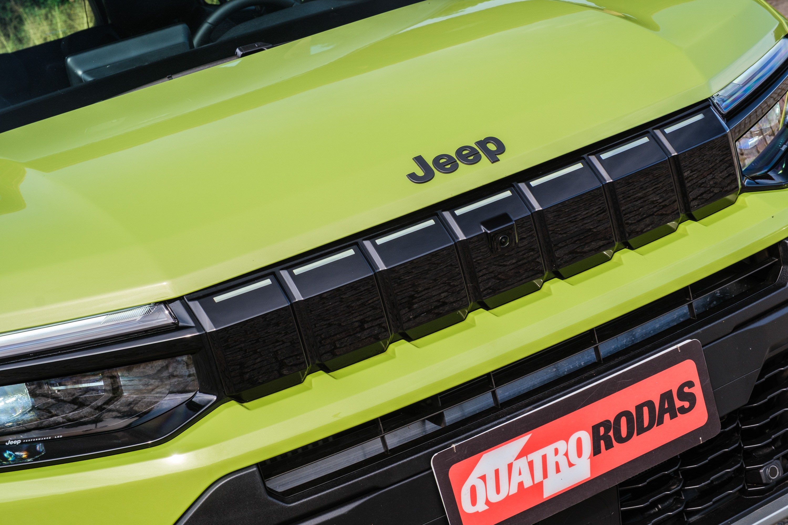 Cap&ocirc; verde-lim&atilde;o de um carro Jeep, com o nome da marca em preto. A grade frontal preta tem sete aberturas retangulares e um farol esquerdo vis&iacute;vel. Abaixo, uma placa vermelha com QUATRO RODAS em branco