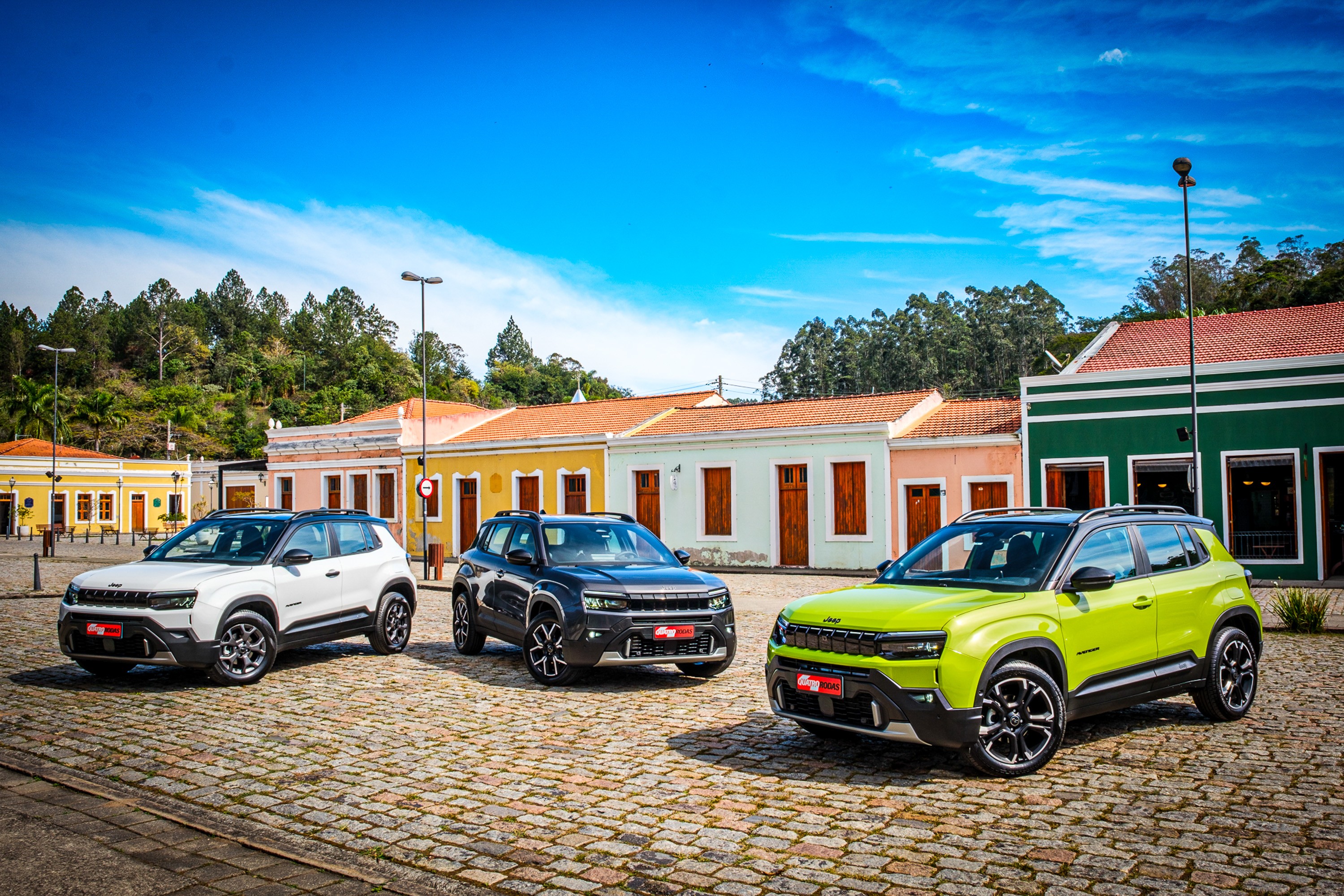 Tr&ecirc;s carros Jeep Avenger estacionados em uma rua de paralelep&iacute;pedos, com casas coloridas ao fundo e um c&eacute;u azul com nuvens esparsas. Da esquerda para a direita, os carros s&atilde;o branco, cinza escuro e verde lim&atilde;o.