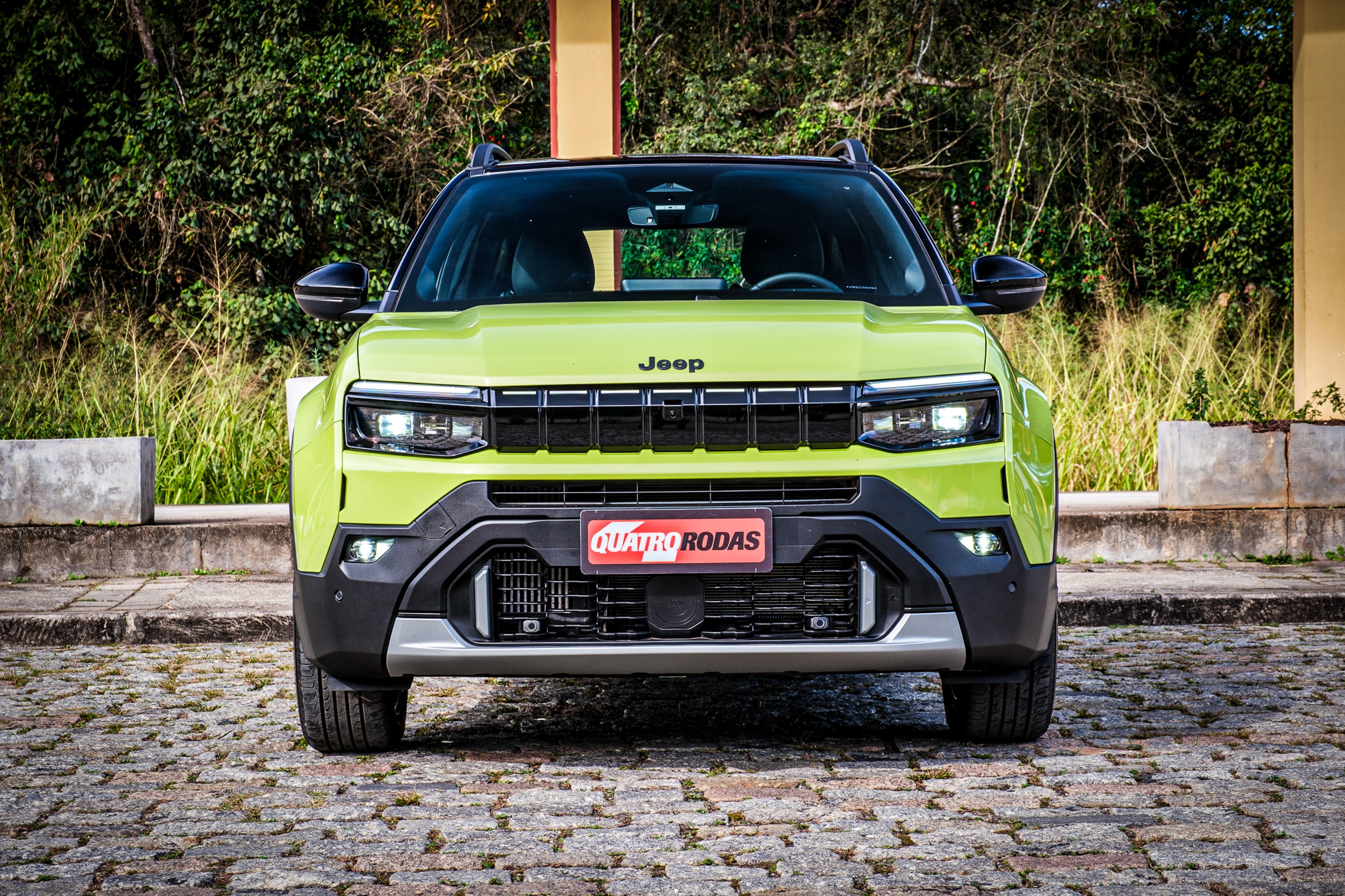 SUV Jeep Avenger verde-lim&atilde;o, visto de frente, estacionado em piso de paralelep&iacute;pedos. O carro tem far&oacute;is retangulares, grade preta com o nome Jeep acima, e para-choque robusto com detalhes em cinza. Uma placa vermelha Quatro Rodas est&aacute; no centro. Ao fundo, vegeta&ccedil;&atilde;o densa e um pilar bege.