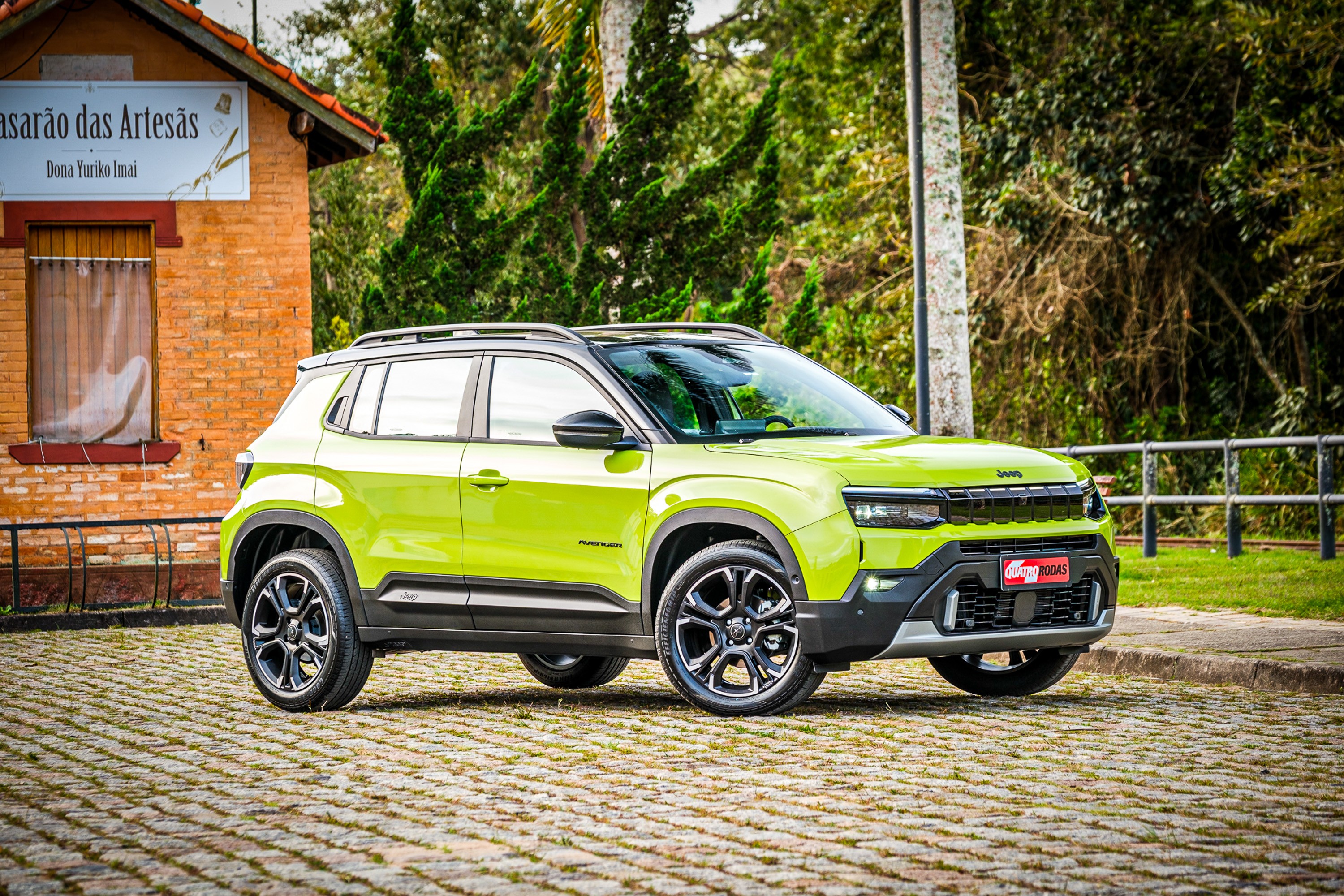 SUV Jeep Avenger verde-lim&atilde;o estacionado em piso de paralelep&iacute;pedos, com uma casa de tijolos &agrave; esquerda e &aacute;rvores ao fundo