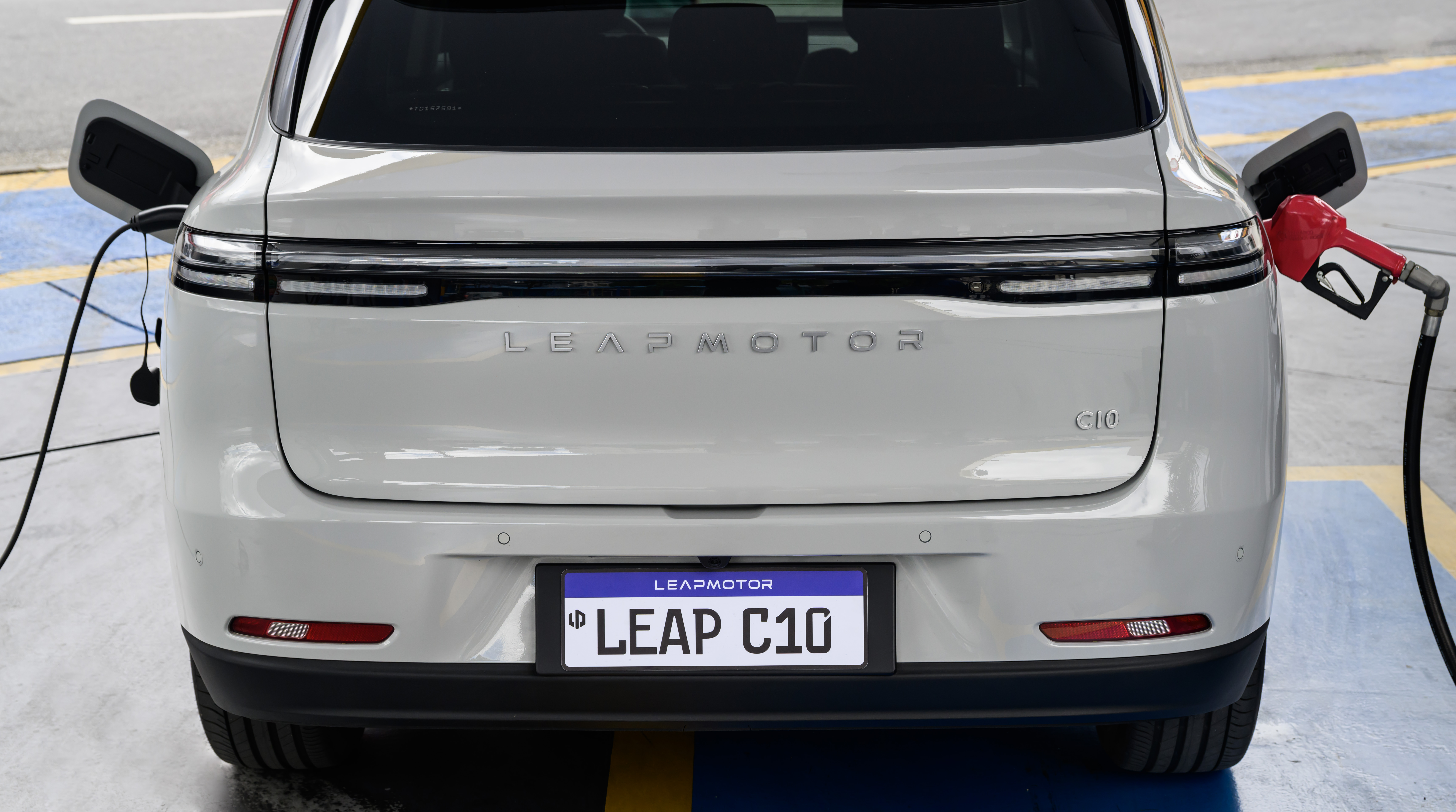 Traseira de um carro branco Leapmotor C10, com um cabo de carregamento el&eacute;trico conectado &agrave; esquerda e um bico de bomba de combust&iacute;vel vermelho inserido &agrave; direita, simbolizando a transi&ccedil;&atilde;o energ&eacute;tica. A placa exibe LEAP C10