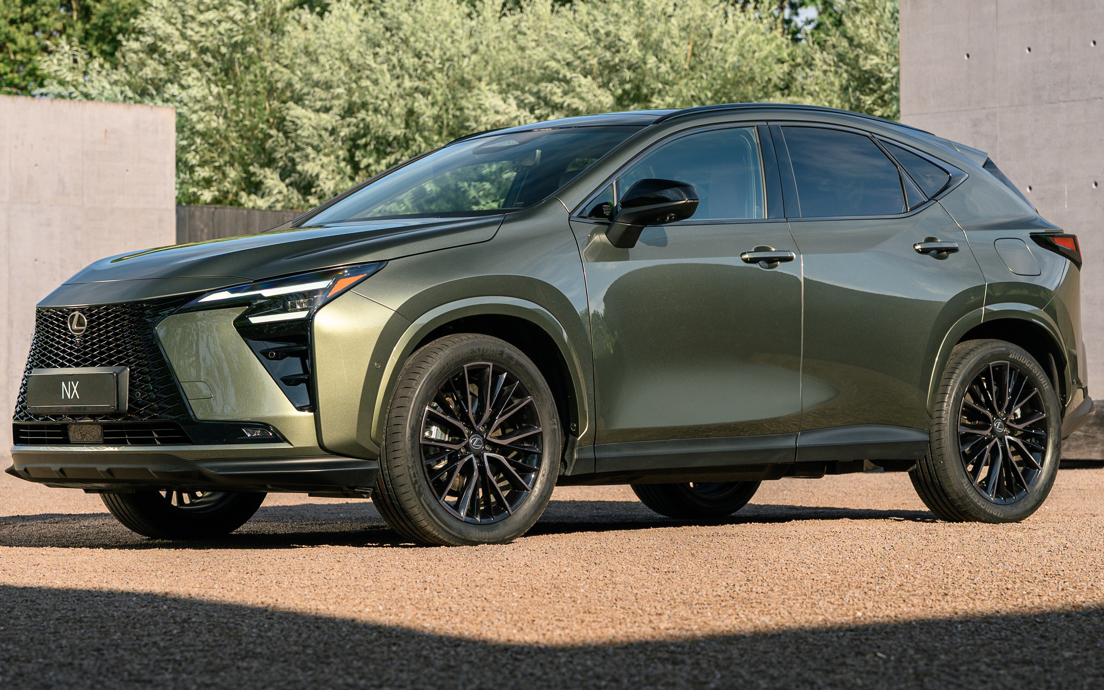 SUV Lexus NX 2024 verde-oliva met&aacute;lico, estacionado em uma &aacute;rea externa com piso de cascalho e vegeta&ccedil;&atilde;o ao fundo. O carro tem rodas pretas e detalhes cromados na grade frontal e far&oacute;is. A luz do sol incide sobre o ve&iacute;culo, criando sombras no ch&atilde;o
