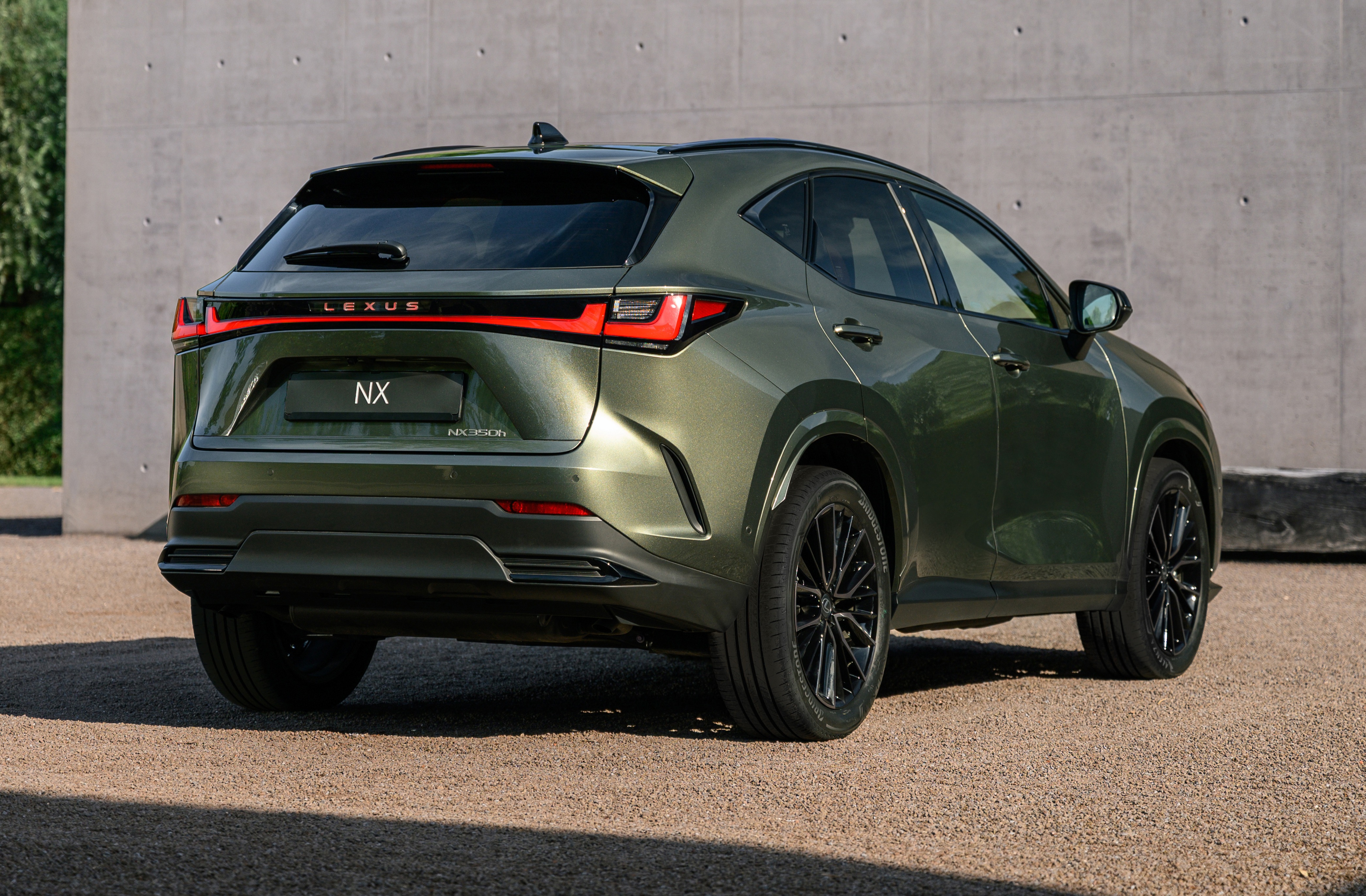 Traseira de um SUV Lexus NX verde-oliva met&aacute;lico, estacionado em uma superf&iacute;cie de cascalho. As lanternas traseiras em LED formam uma linha horizontal vermelha, com o nome LEXUS acima e NX no centro da tampa do porta-malas. Rodas pretas com m&uacute;ltiplos raios e detalhes cromados. Ao fundo, uma parede de concreto cinza e vegeta&ccedil;&atilde;o verde &agrave; esquerda