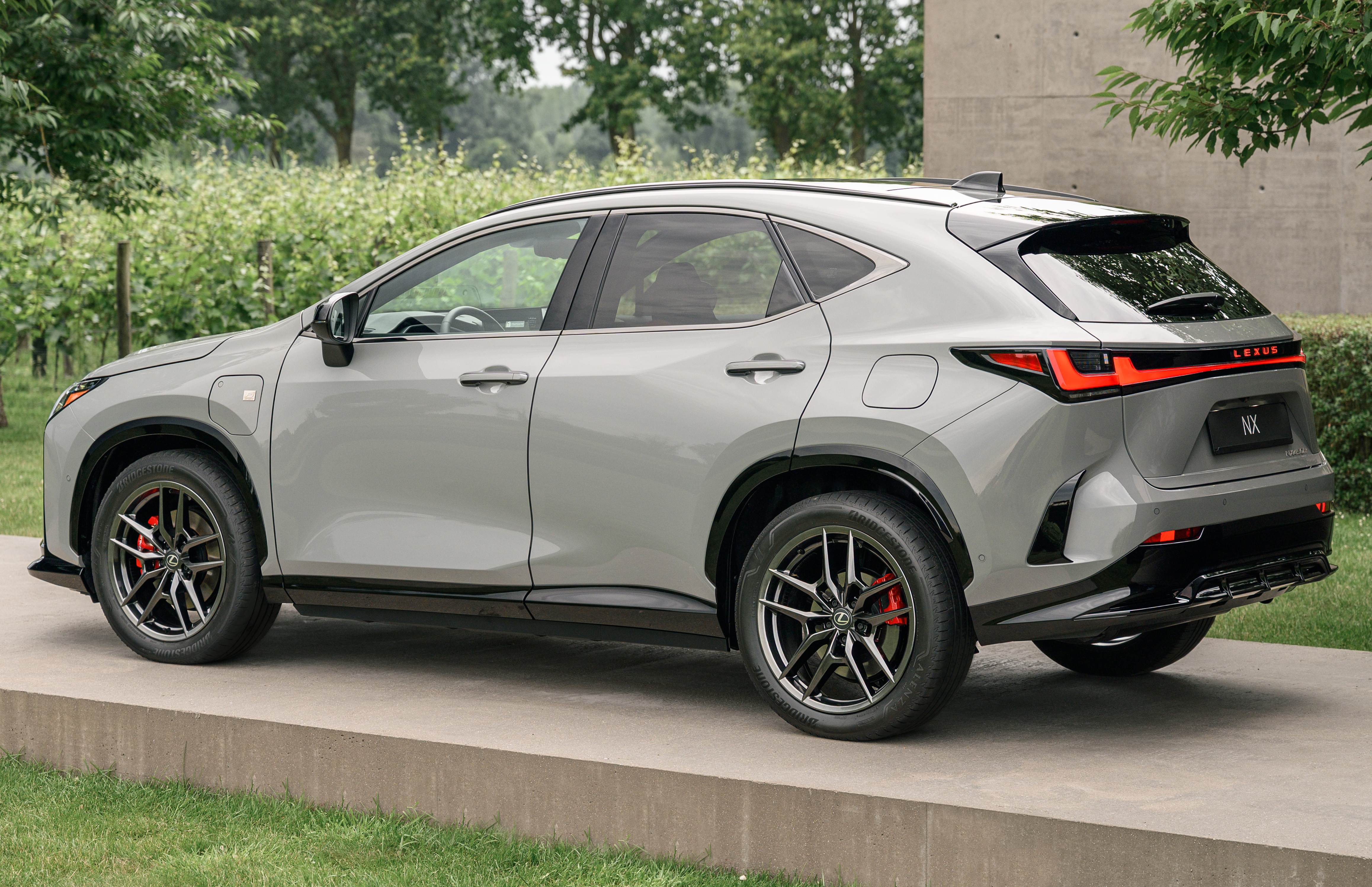 SUV Lexus NX cinza claro, visto de lado, estacionado em uma plataforma de concreto com grama ao redor. O carro tem detalhes pretos nas rodas e na parte inferior da carroceria, e lanternas traseiras em LED vermelhas e pretas. Ao fundo, &aacute;rvores e vegeta&ccedil;&atilde;o verde