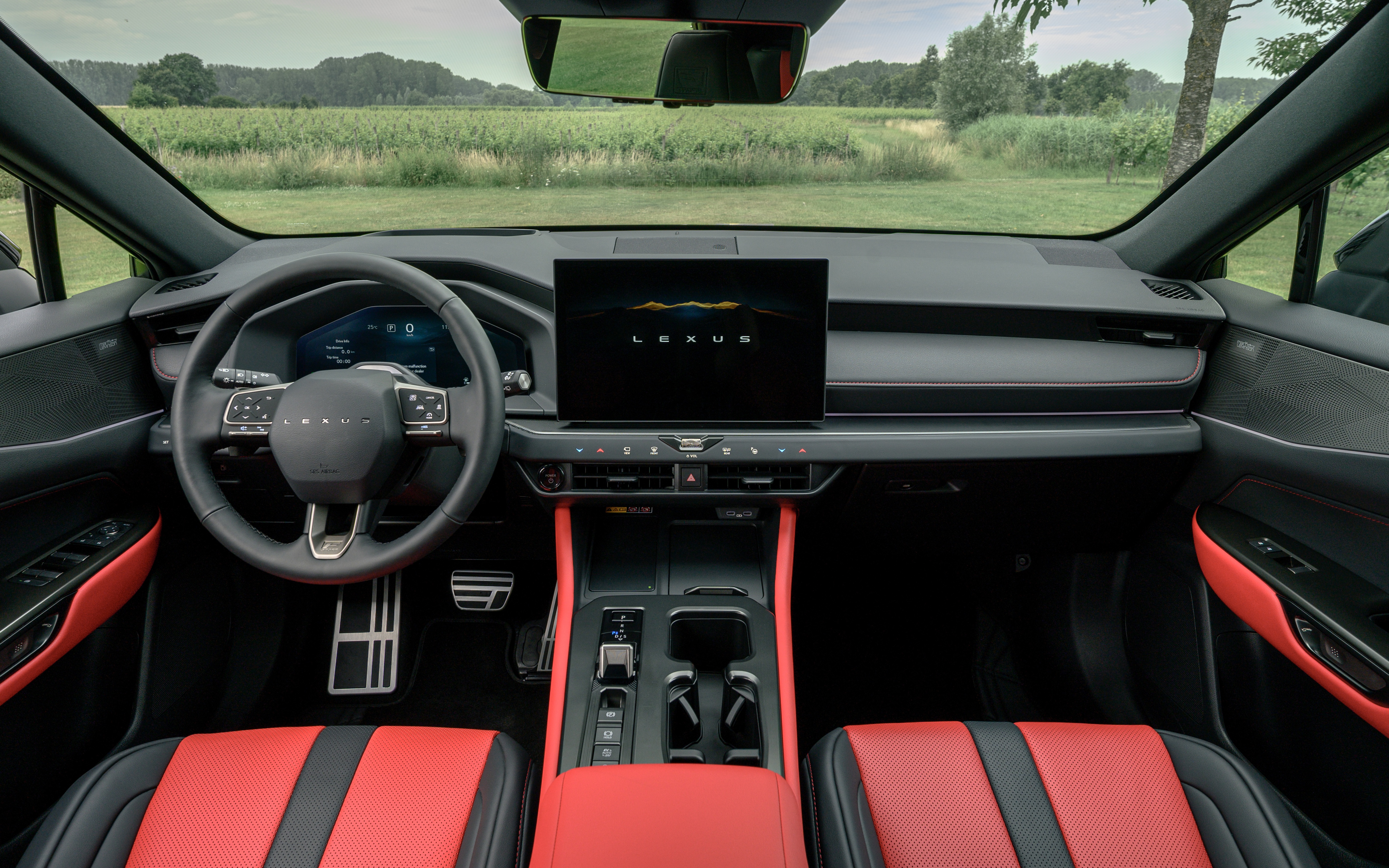 Interior de um carro Lexus com bancos esportivos vermelhos e pretos, volante multifuncional escuro e painel com tela central exibindo o logo Lexus. O para-brisa revela um campo verde com &aacute;rvores ao fundo sob um c&eacute;u claro