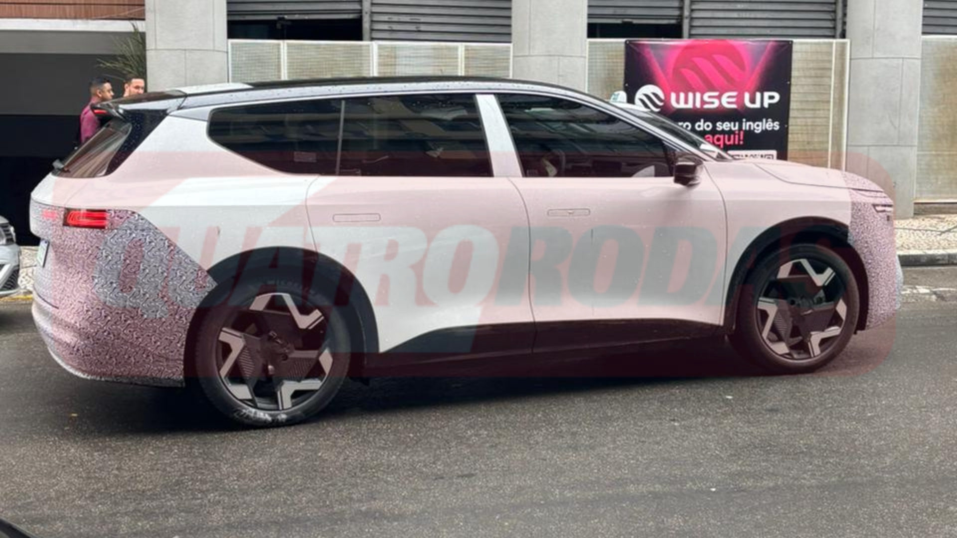 Um carro SUV branco com camuflagem cinza e preta nas extremidades, estacionado na rua. Ao fundo, um banner da Wise Up e duas pessoas conversando