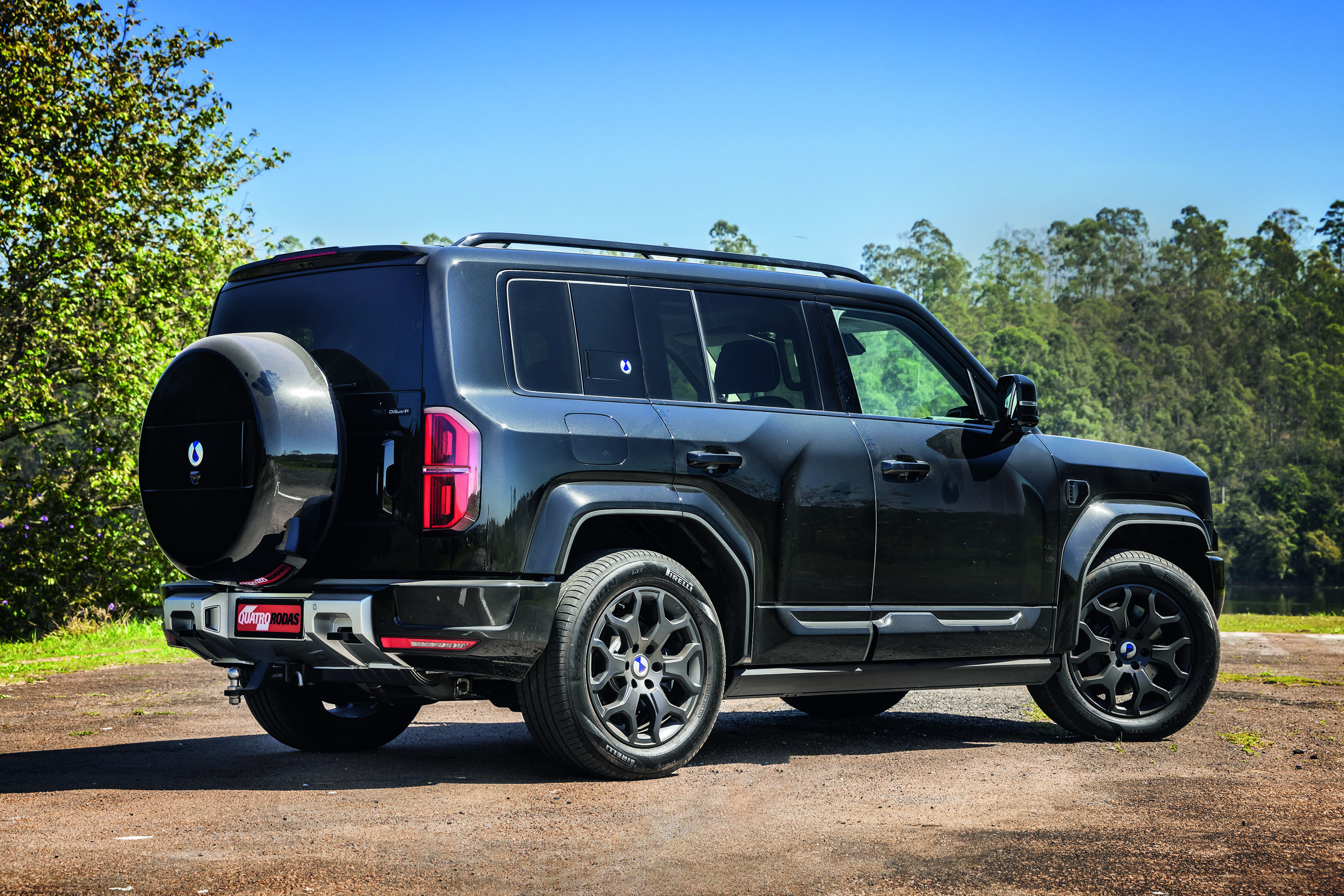 SUV preto brilhante estacionado em uma estrada de terra, com a traseira e lateral direita vis&iacute;veis. Possui estepe na tampa traseira, rack de teto e rodas escuras com design complexo. Ao fundo, c&eacute;u azul claro e &aacute;rvores verdes
