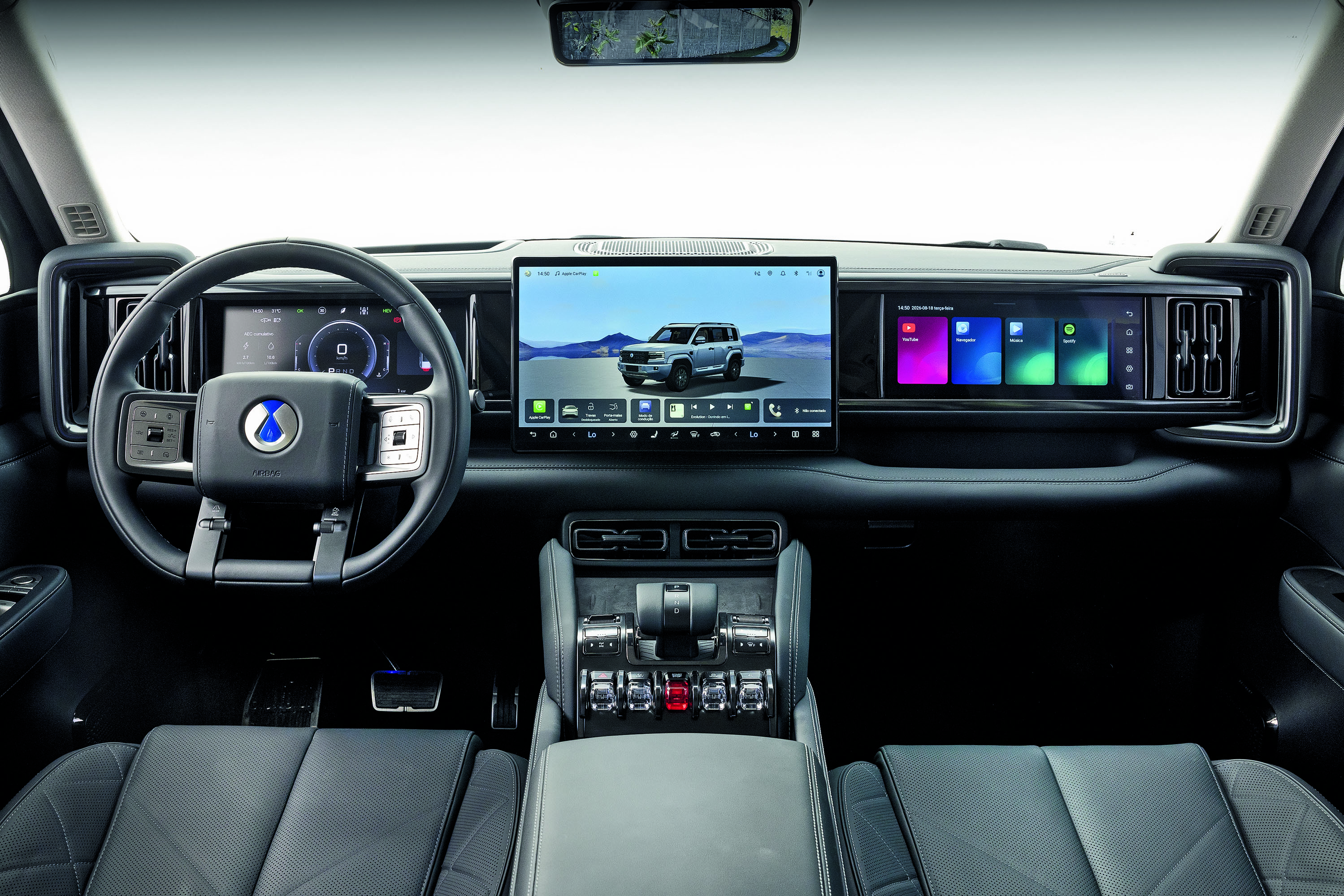 Interior de um carro moderno com painel digital. O volante preto tem um logo azul no centro. &Agrave; direita, uma tela exibe um SUV cinza em paisagem montanhosa. Outra tela menor mostra aplicativos coloridos. Bancos de couro pretos e console central com bot&otilde;es prateados completam o design.
