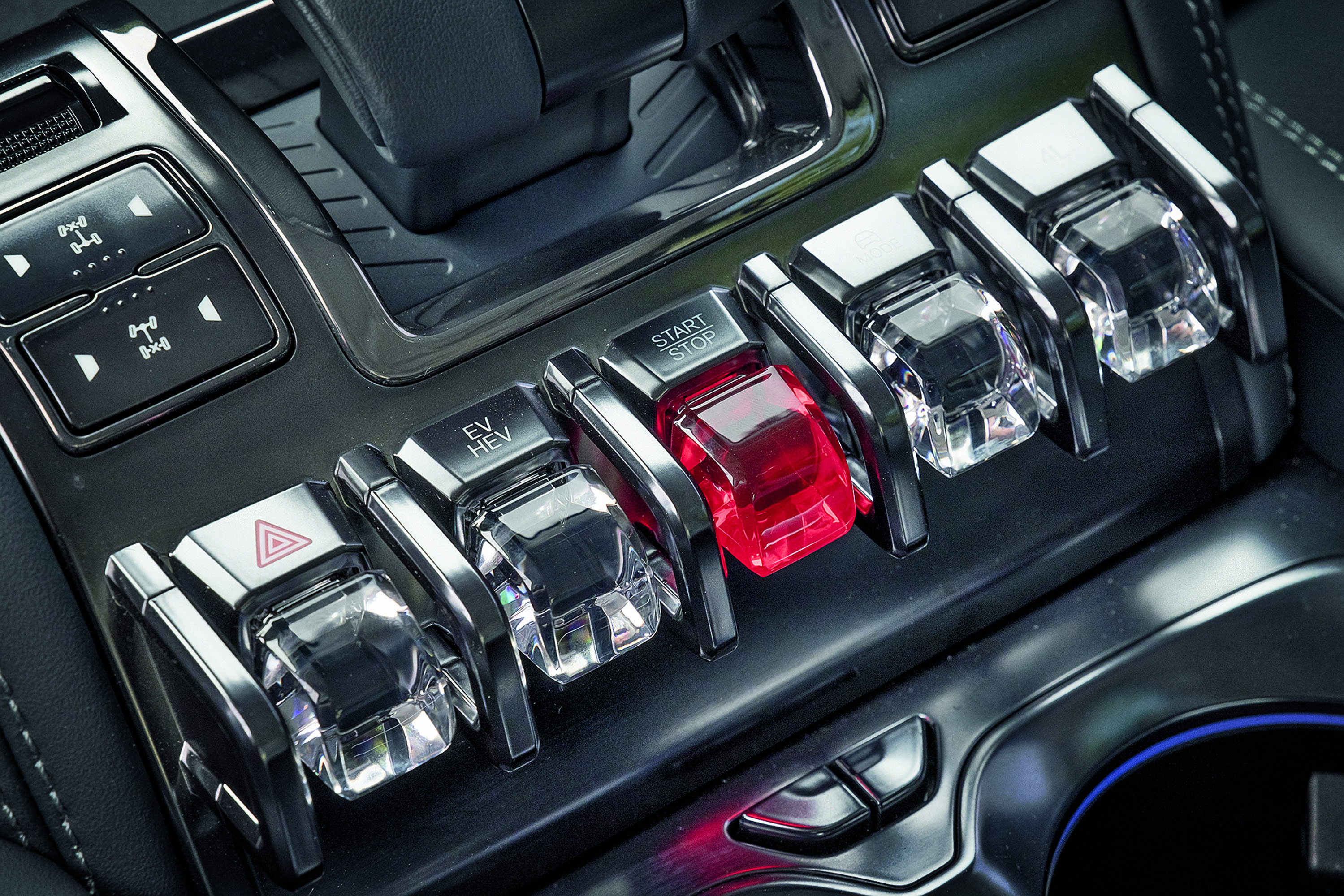 Painel de controle de carro com cinco bot&otilde;es tipo alavanca, o central vermelho com START STOP, e os outros transparentes com s&iacute;mbolos de alerta, tra&ccedil;&atilde;o e modo el&eacute;trico
