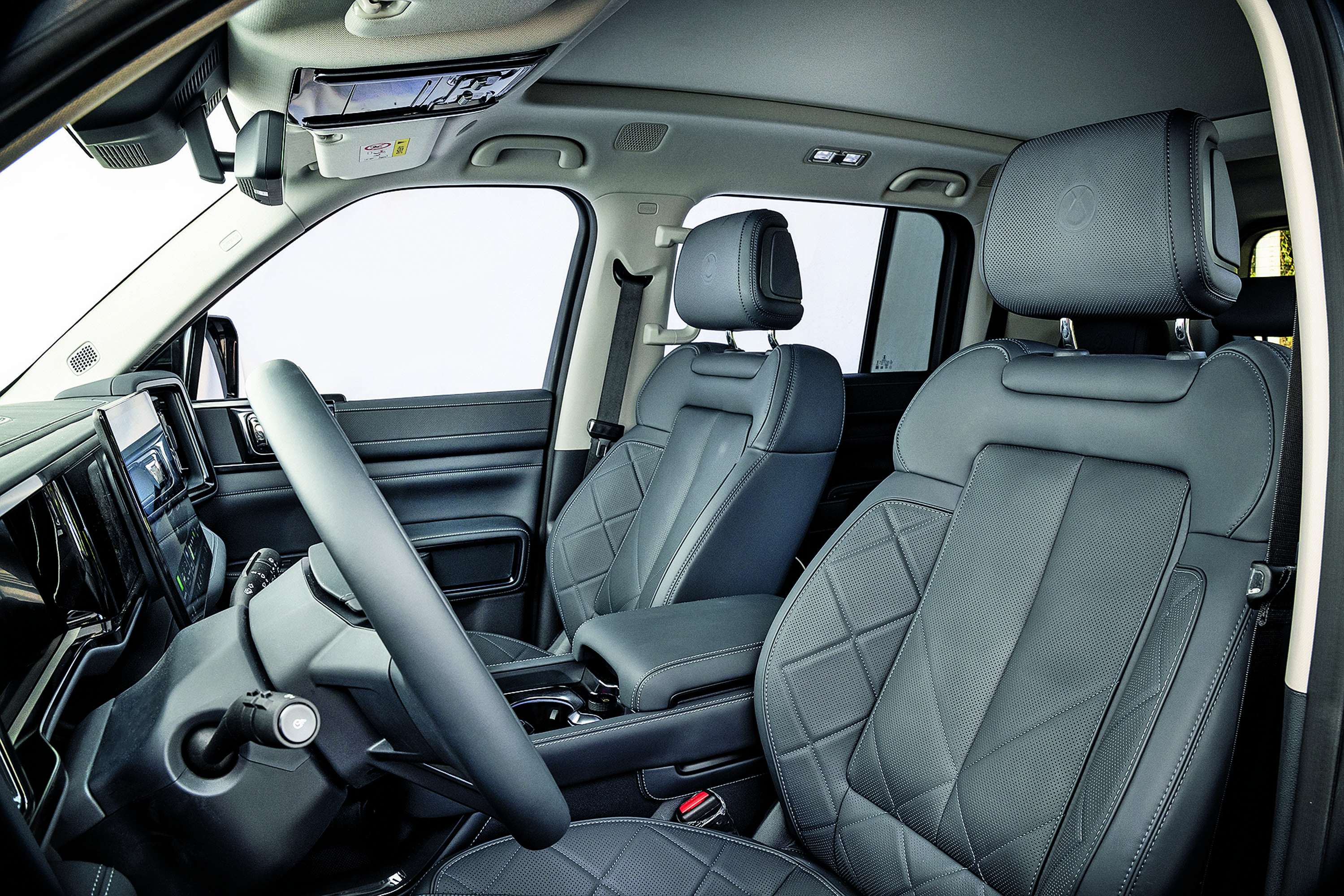 Interior de um carro com bancos de couro cinza escuro e costuras em losango, volante &agrave; esquerda e painel digital. O banco do motorista e o do passageiro dianteiro s&atilde;o vis&iacute;veis, com o banco traseiro parcialmente &agrave; direita. O teto solar e o espelho retrovisor interno tamb&eacute;m aparecem