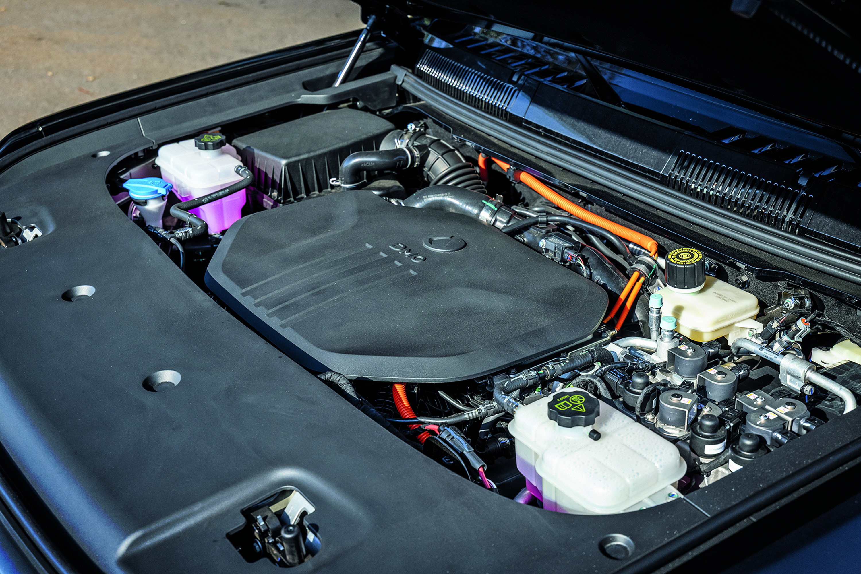 Compartimento do motor de um carro h&iacute;brido ou el&eacute;trico, com cabos laranja de alta voltagem, reservat&oacute;rios de fluidos e uma tampa preta cobrindo parte do motor