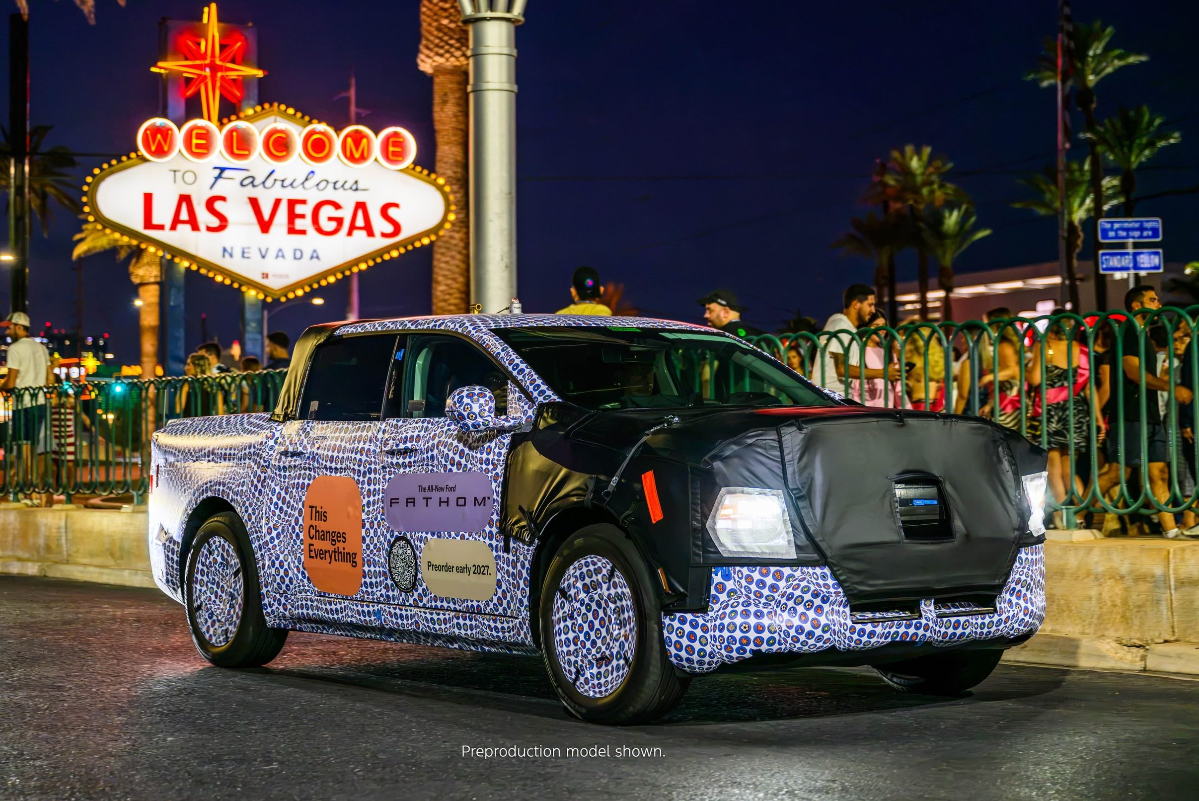 Picape camuflada com padrão azul e branco, com a frente coberta por tecido preto, estacionada à noite em Las Vegas, Nevada. Ao fundo, o letreiro luminoso Welcome to Fabulous Las Vegas e palmeiras iluminadas. Pessoas e grades verdes aparecem no lado direito da imagem
